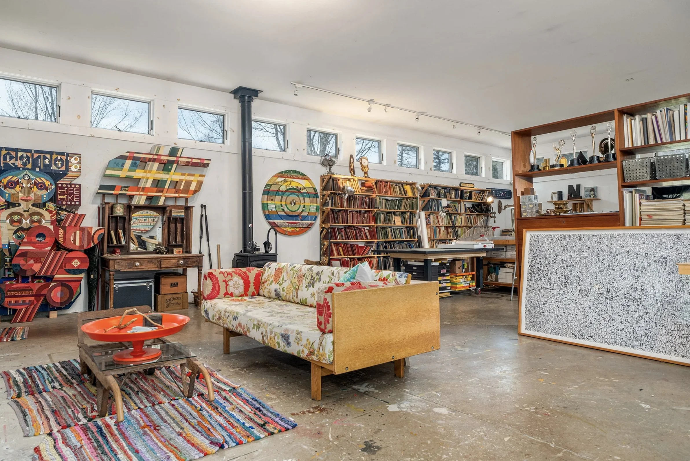 Interior of an art studio or library with colorful abstract artwork on the walls, a vintage sofa with patterned upholstery, a small wooden coffee table with a red dish, a multicolored woven rug, and shelves filled with books and trophies.