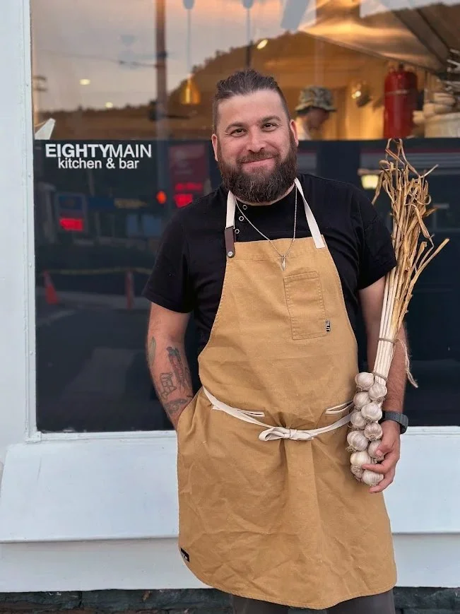 Man with a beard wearing a black t-shirt and an apron holding a bunch of garlic bulbs and dried herbs outside of EIGHTYMAIN kitchen & bar establishment.