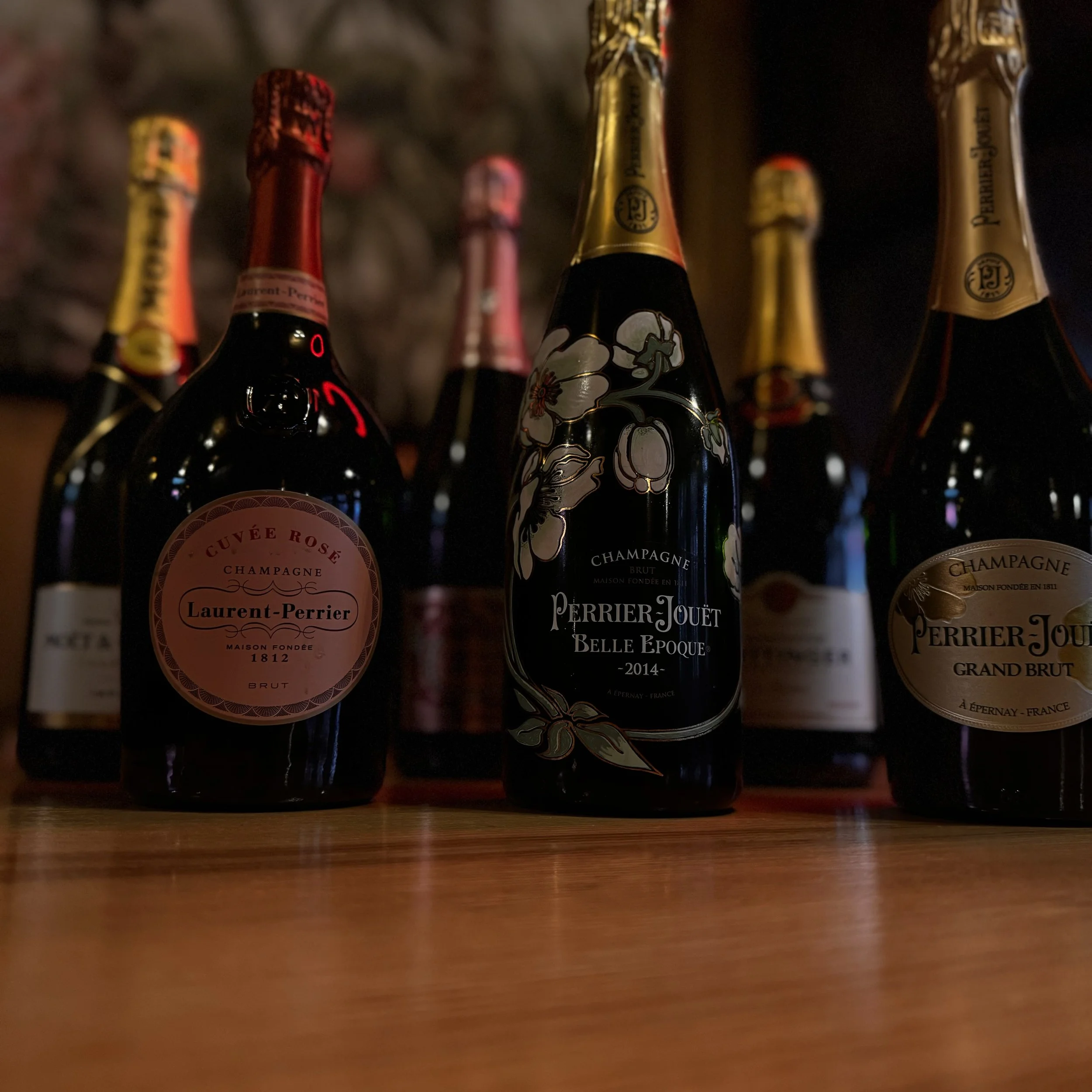 Several bottles of Perrier-Jouët champagne, including Belle Époque, Grand Brut, and Cuvée Rosé, arranged on a wooden surface with a brick wall background.