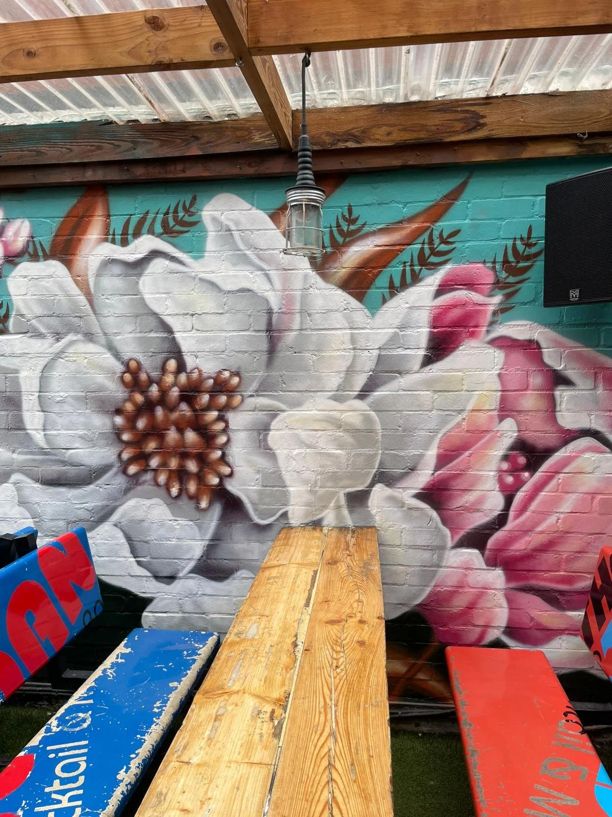 Colorful graffiti mural of a white flower with pink accents and brown center on a brick wall, with wooden roof beams, a hanging lamp, and picnic benches in front.