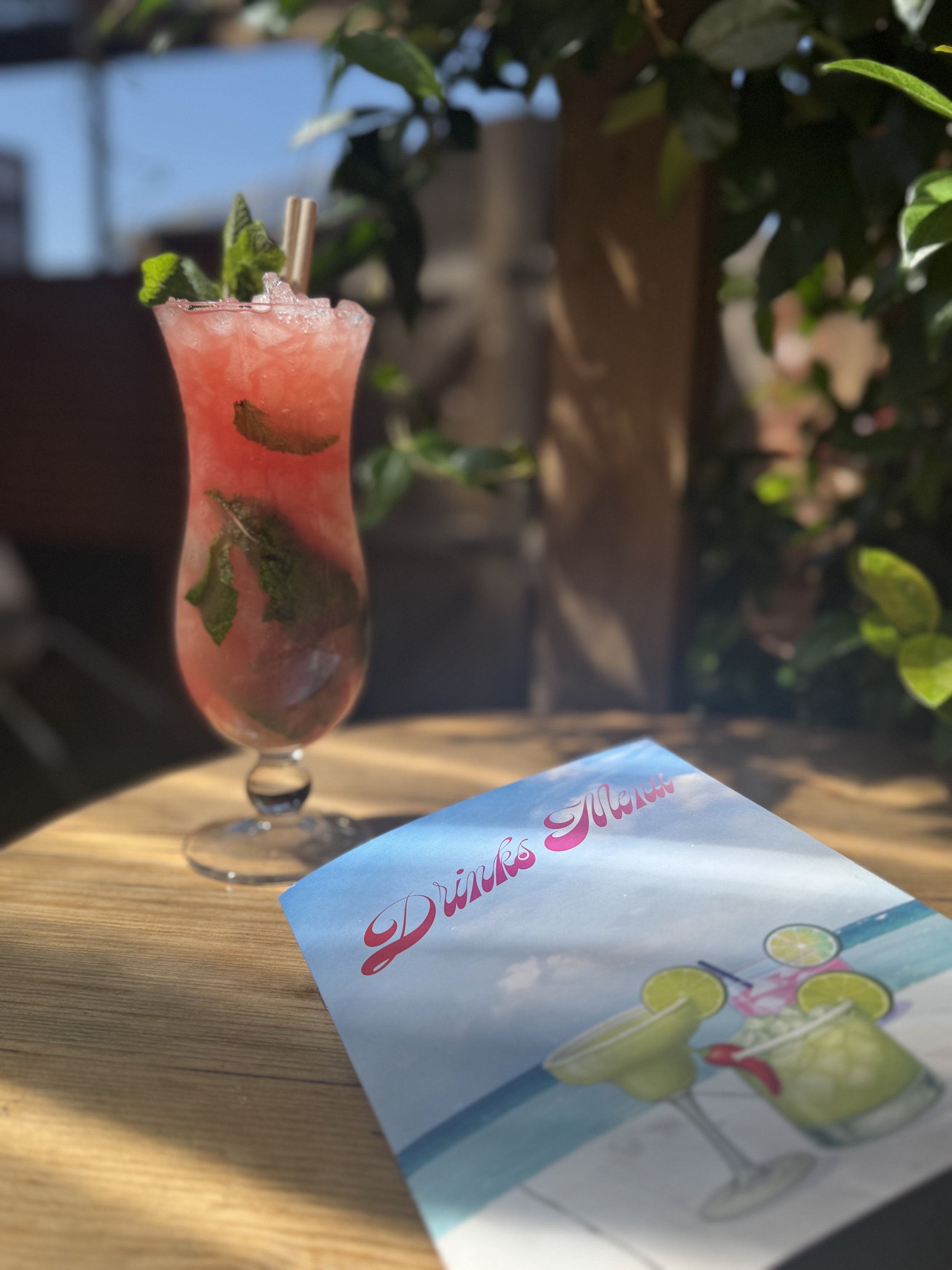 A pink cocktail drink garnished with mint leaves, served in a tall, curvy glass, placed on a wooden table outdoors. A colorful menu titled 'Drinks Menu' with images of cocktails is also on the table.