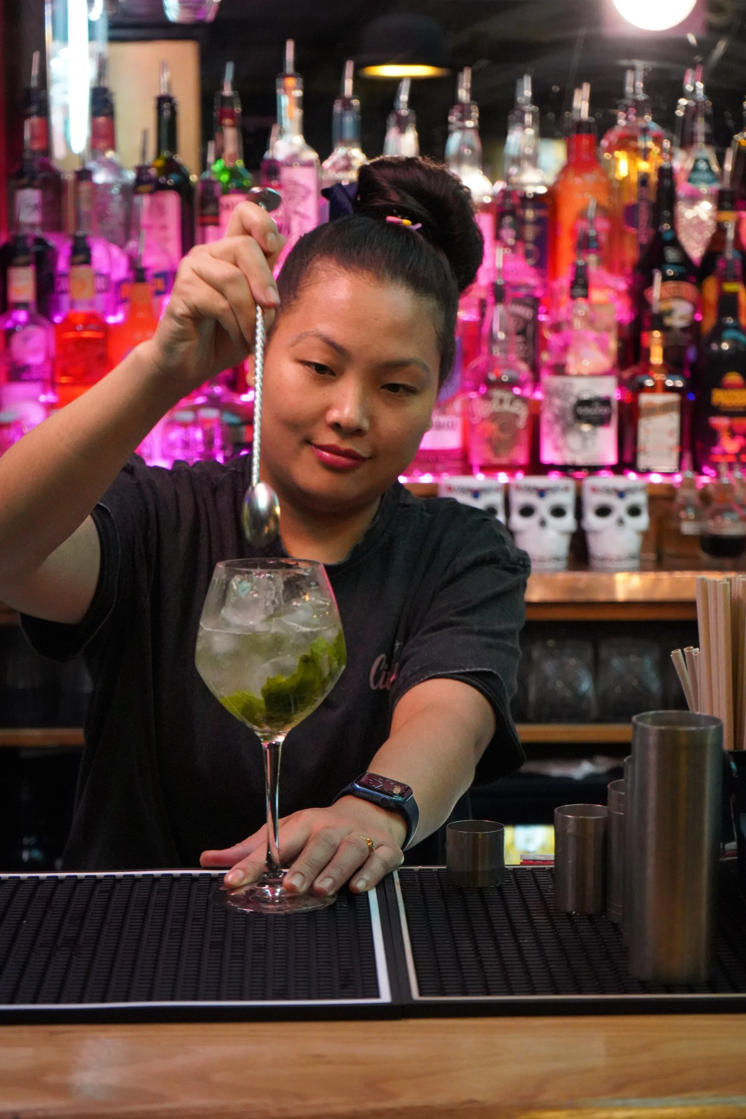 A female bartender preparing a mojito cocktail at a bar with colorful liquor bottles in the background.