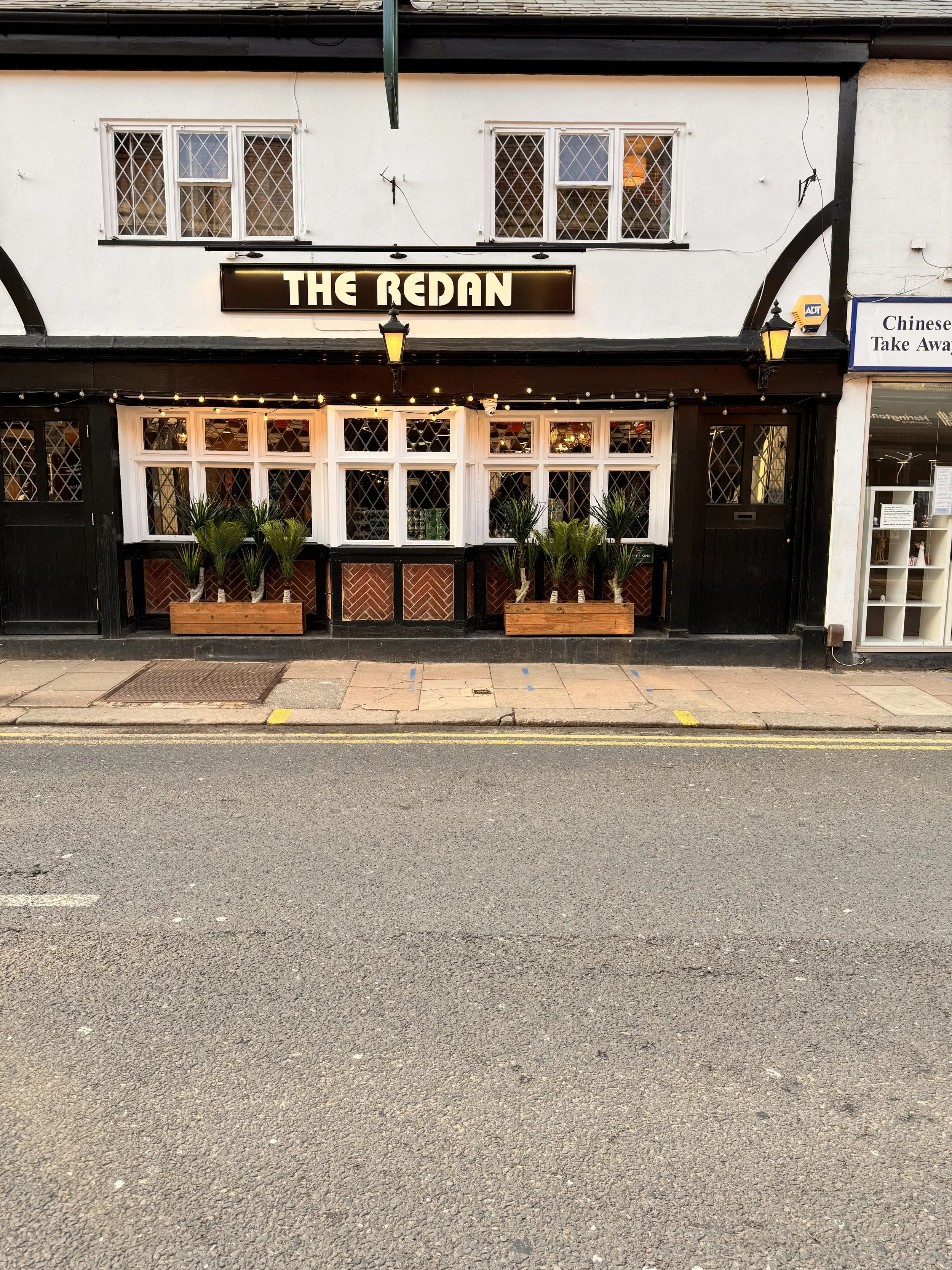 Front of the bar called The Redan with black and white exterior, decorated with string lights and potted palm trees, on a main street in Wokingham.