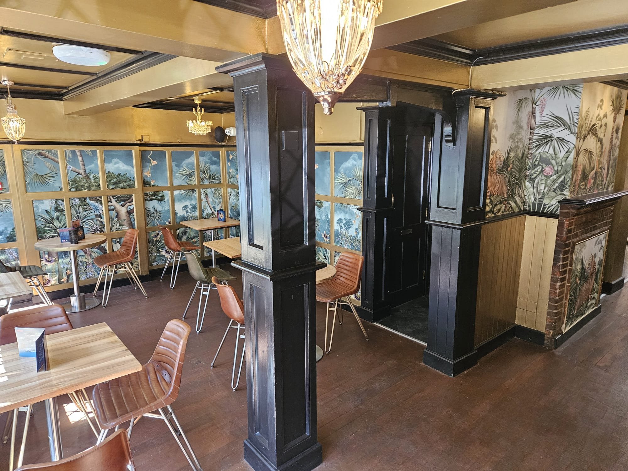 Interior of a restaurant with wooden tables and chairs, dark wood paneling, tropical-themed wallpaper, and a chandelier hanging from the ceiling.