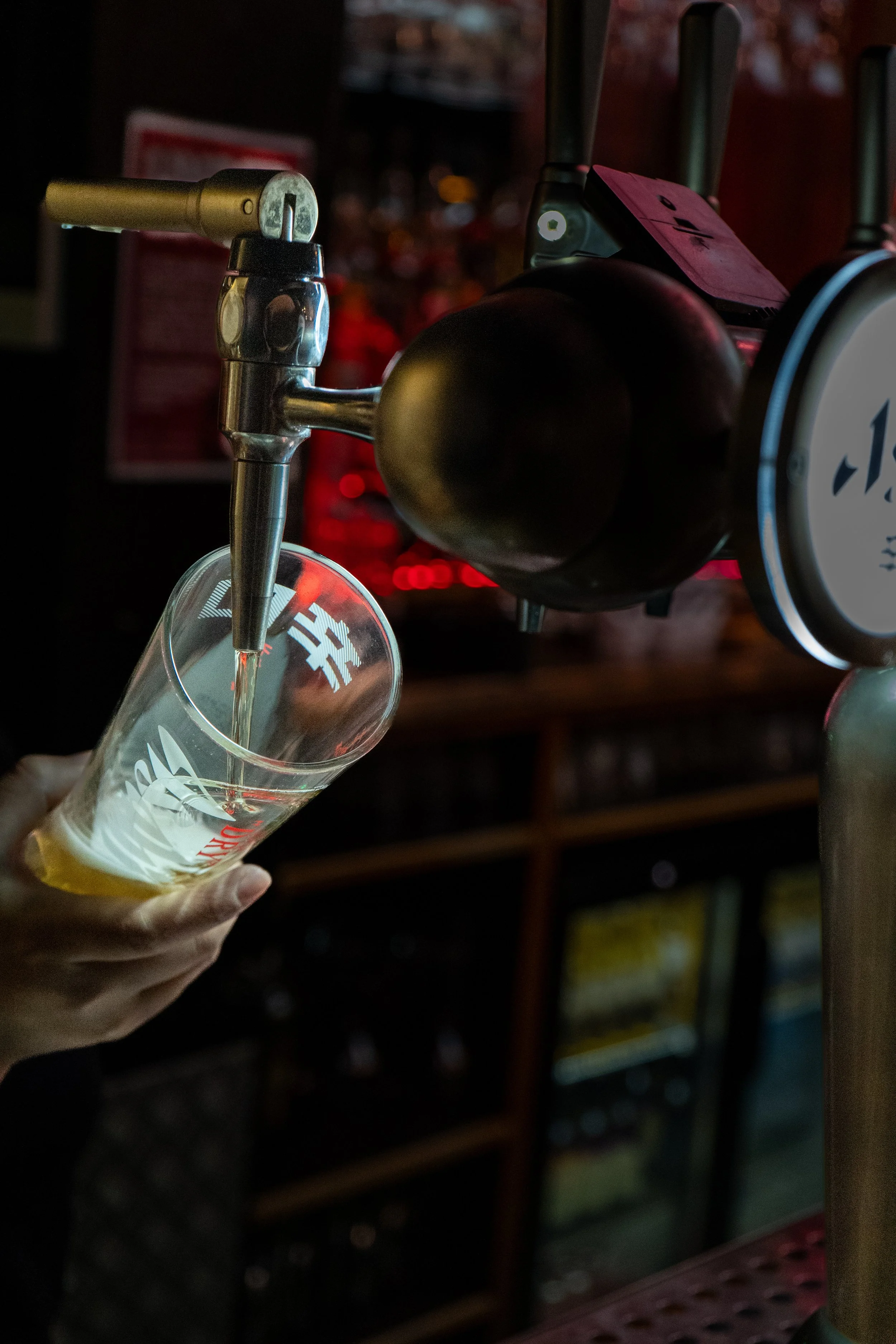 A person pours beer from a tap into a glass at a bar or pub, with a blurred background of shelves and bottles.