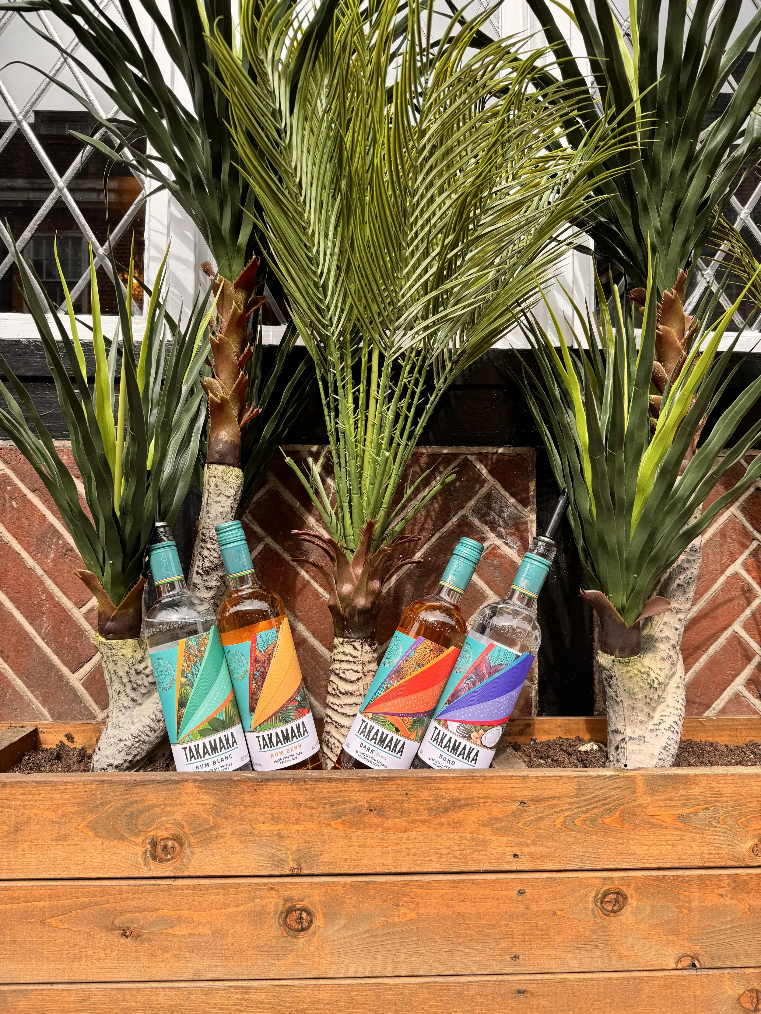 Three bottles of Takamaka rum placed among tall green palm plants with large, broad leaves, in front of a brick wall and window.