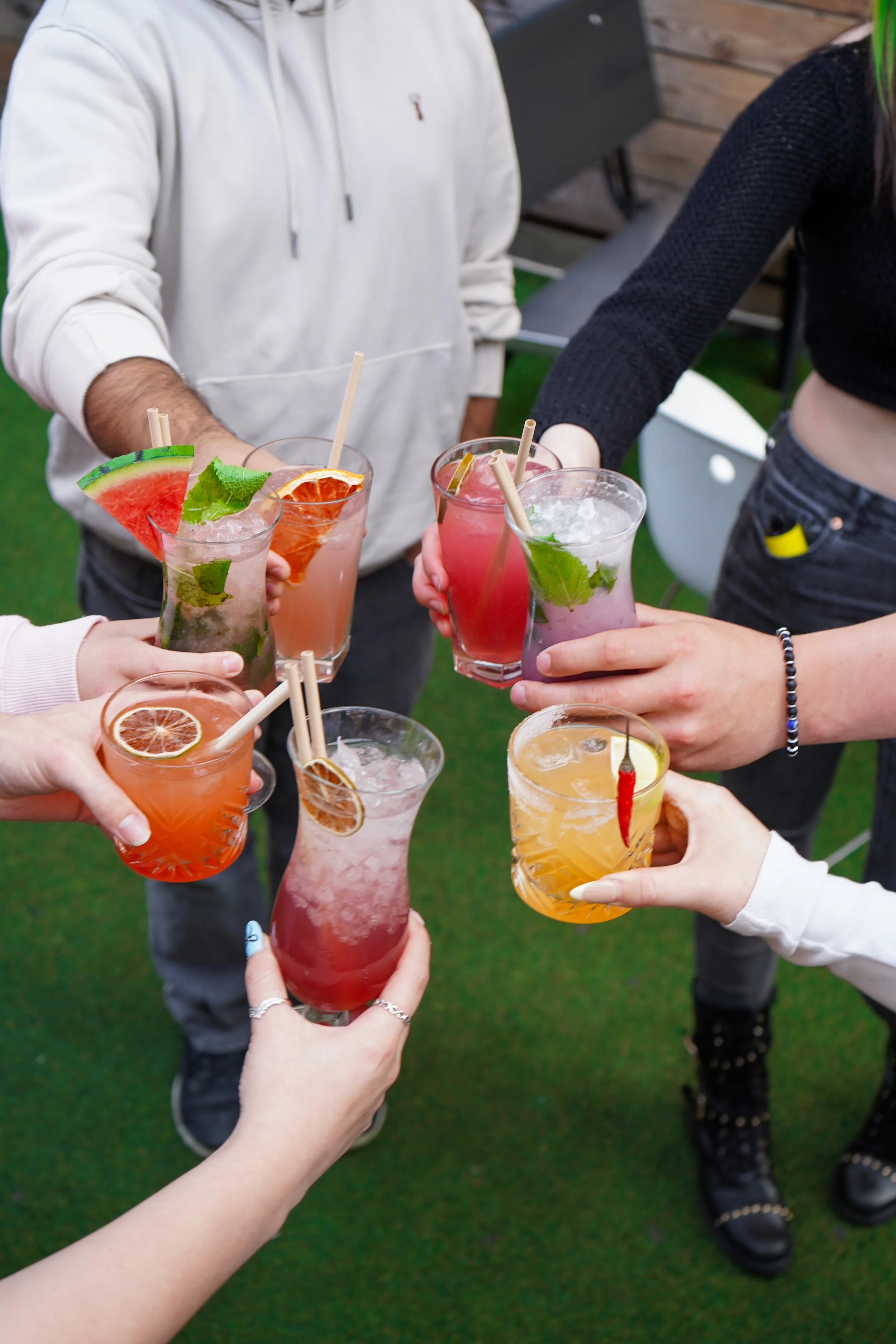 People holding colorful cocktails in glasses with garnishes and straws, celebrating outdoors.