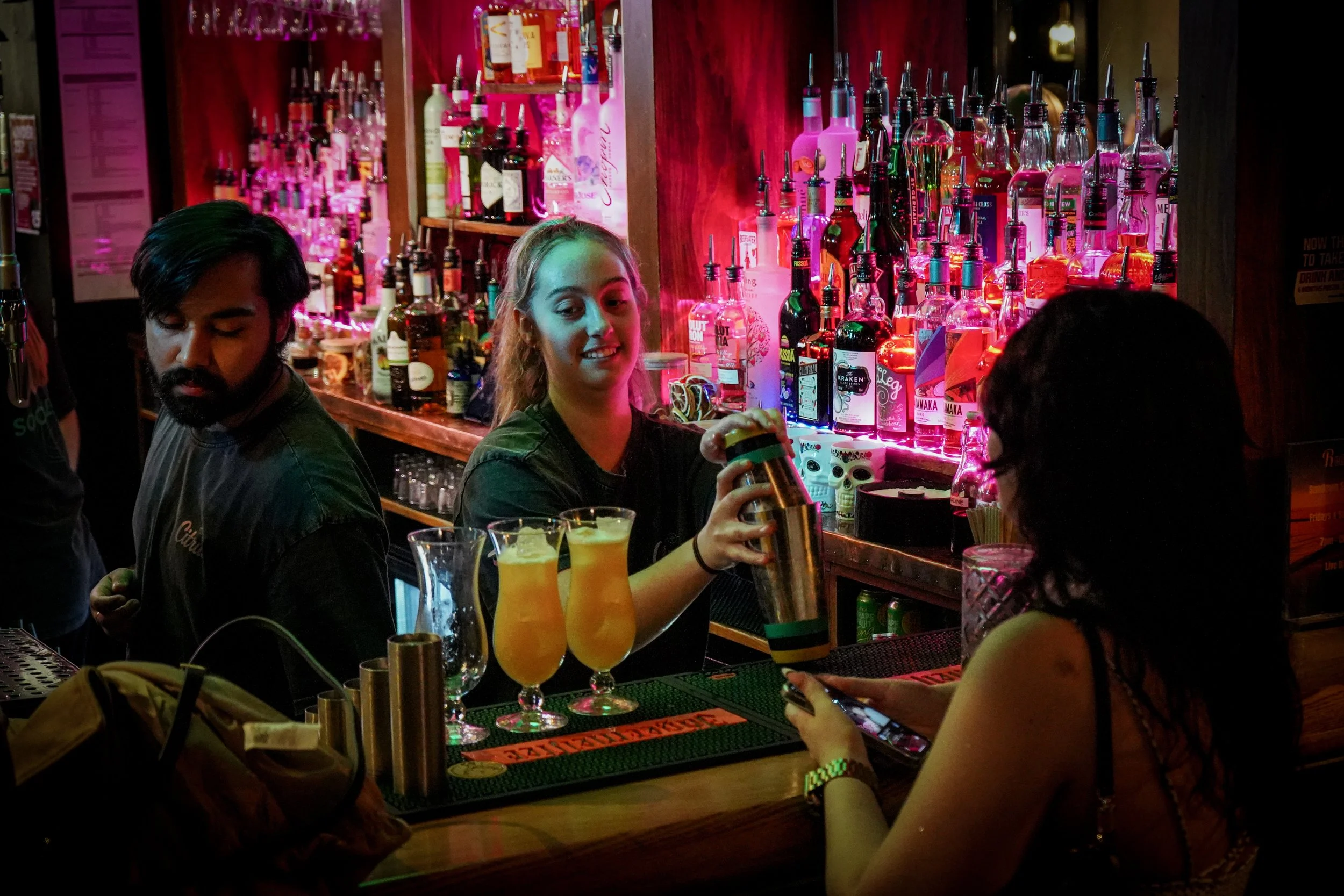 A bartender with blonde hair is preparing a drink for a customer at a bar with colorful liquor bottles in the background, two cocktails on the counter, and a woman using a smartphone.