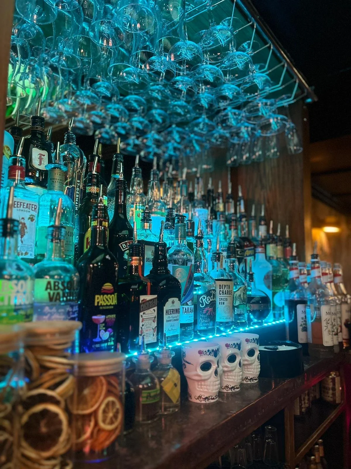 Bar shelf with various liquor bottles and hanging glassware, illuminated with blue lighting, decorated with skull-shaped cups and containers with dried citrus slices.