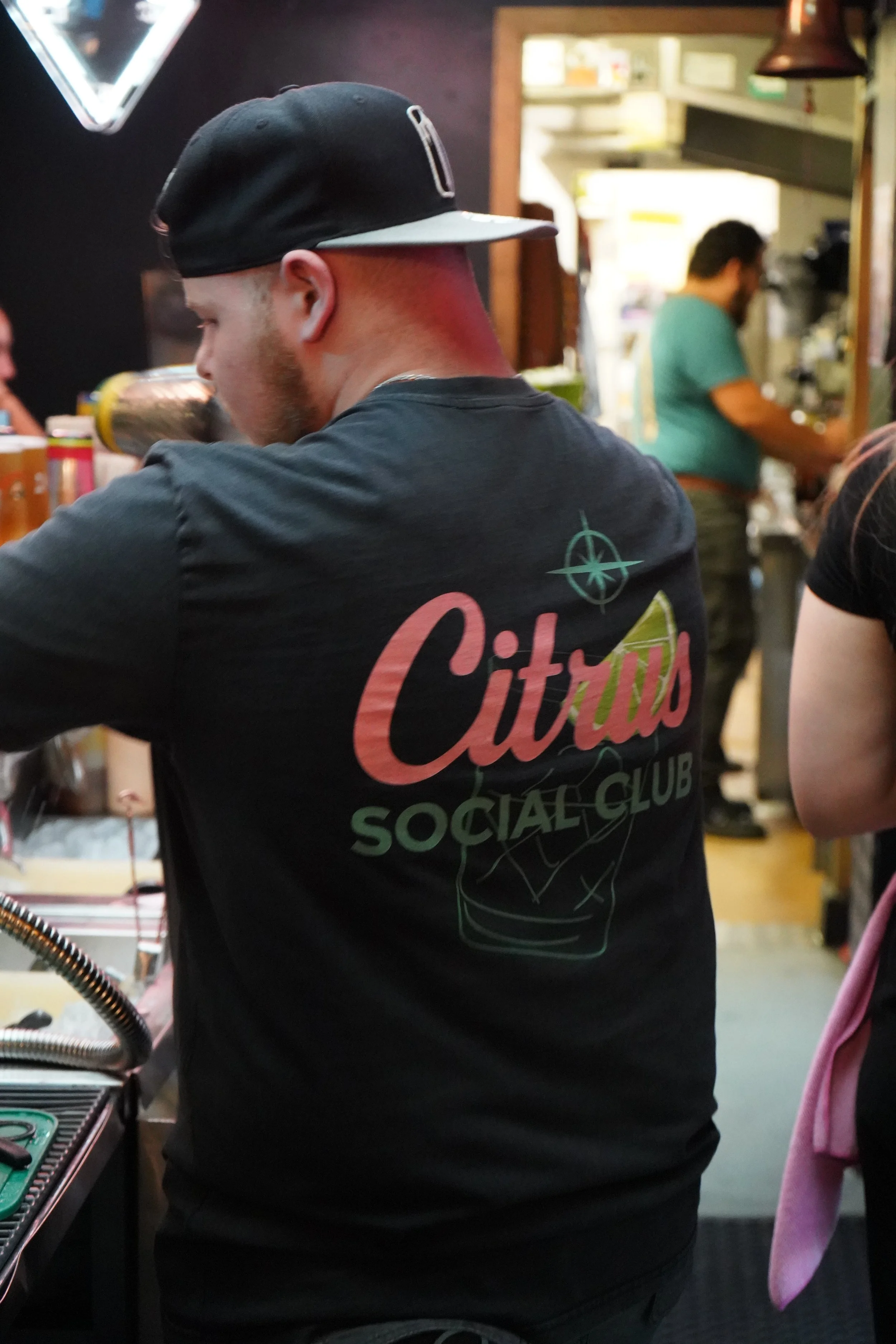 A man wearing a black cap and a 'Citrus Social Club' t-shirt is behind a bar counter, with a kitchen area visible in the background.