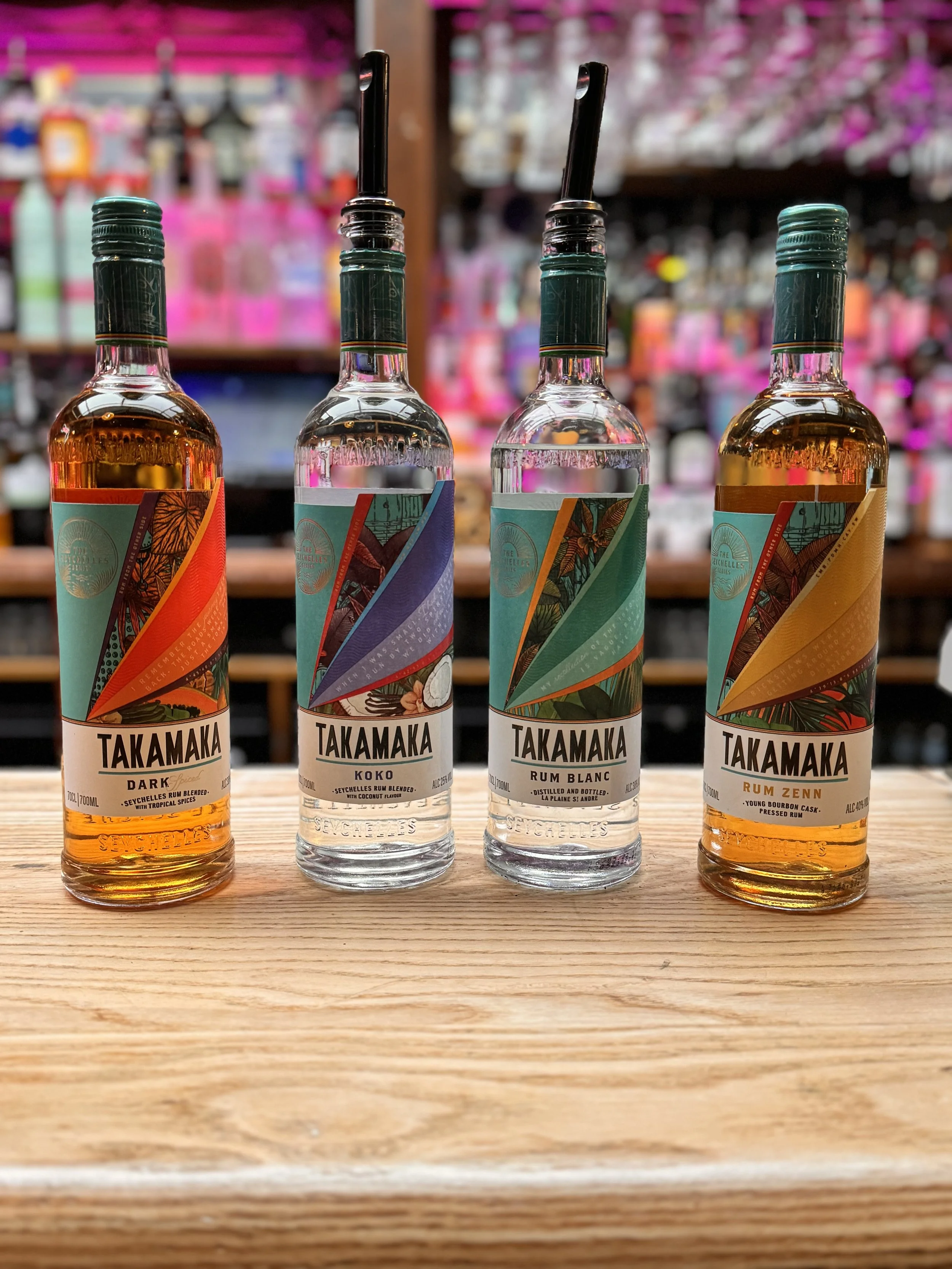 Four bottles of Takamaka rum displayed on a wooden surface with a colorful, blurred store background.