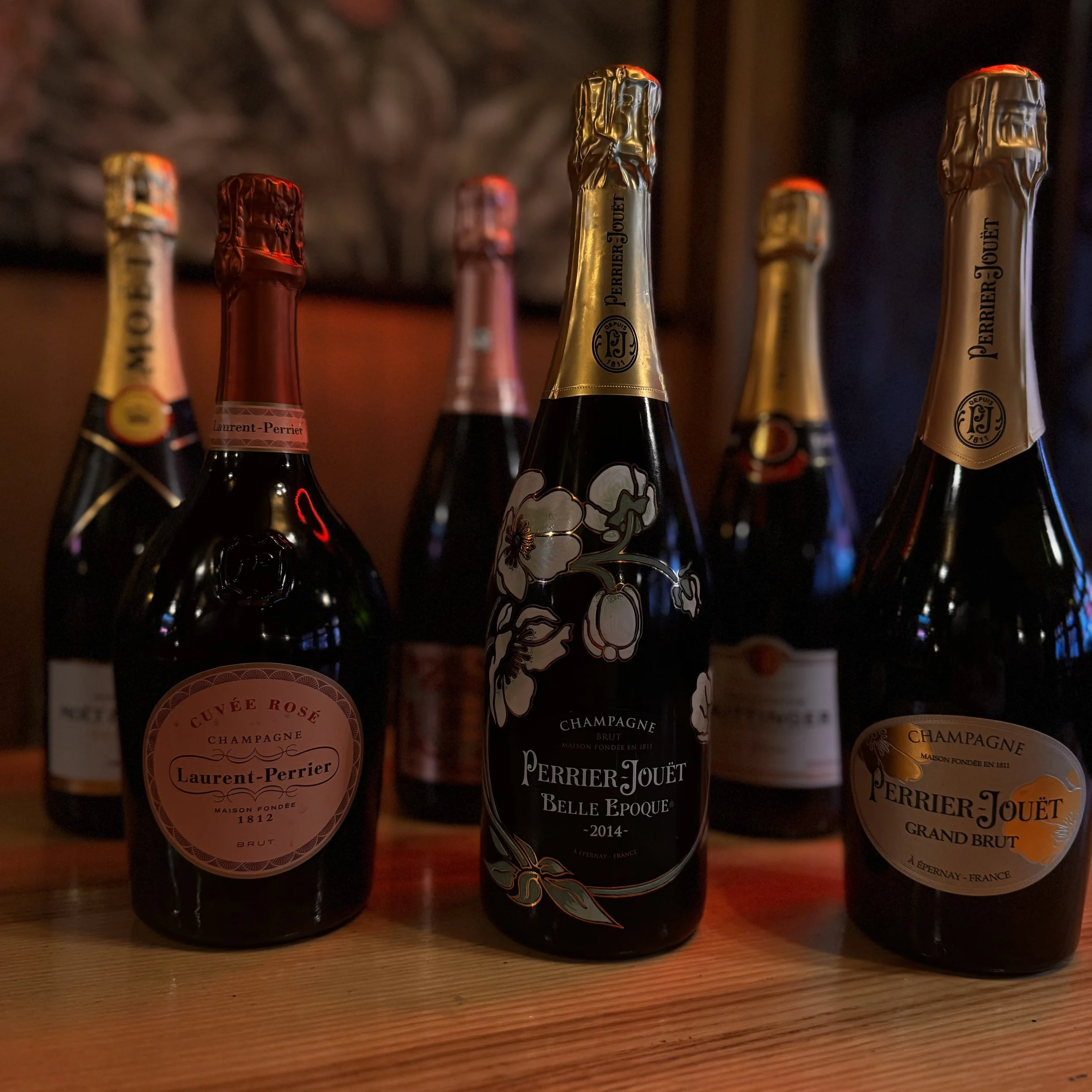Several bottles of Perrier-Jouët champagne, including a Rosé, Belle Époque, and Grand Brut, arranged on a wooden surface in a dimly lit setting.