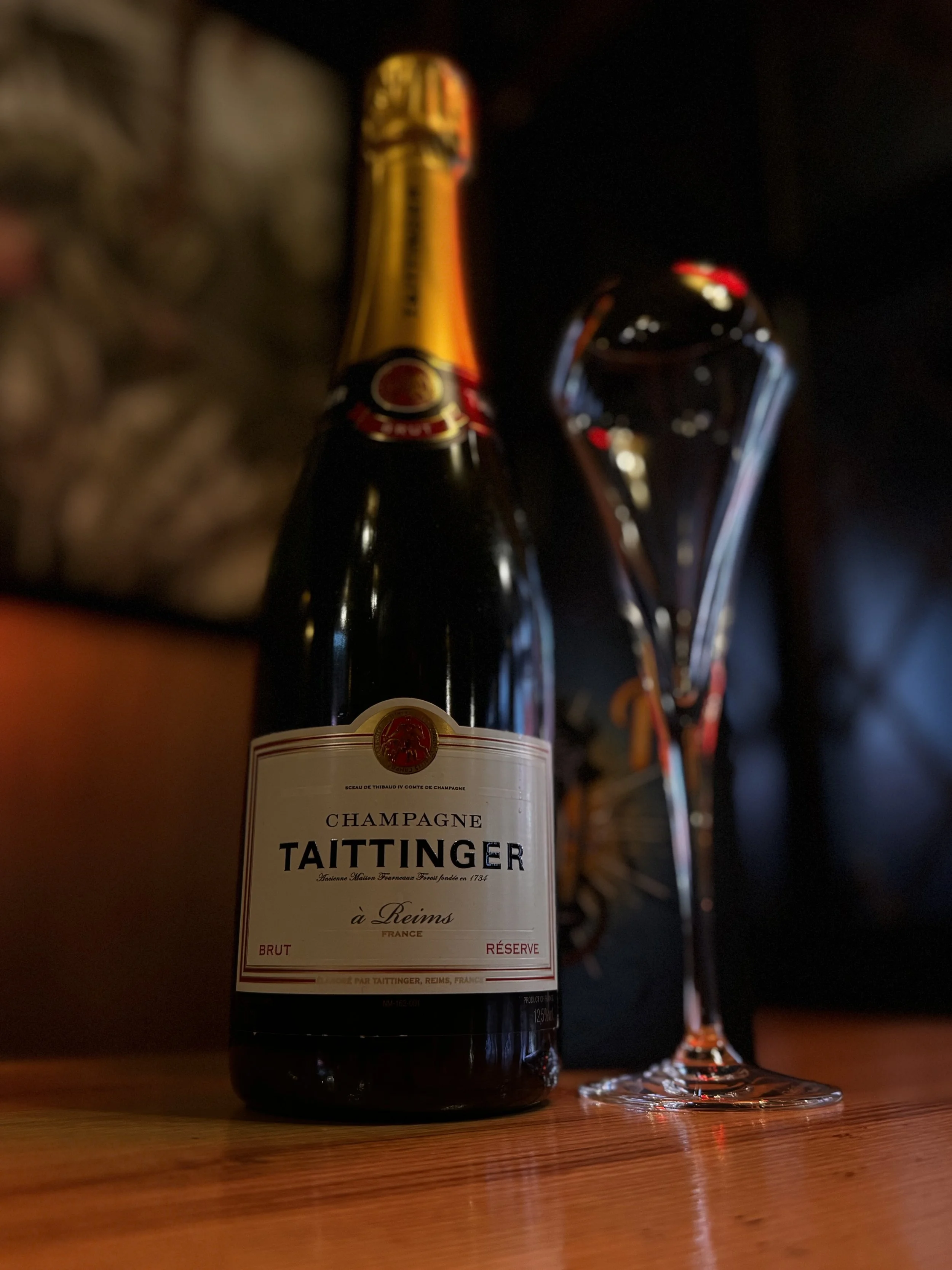 A bottle of Taittinger champagne and a filled glass on a wooden surface with a dark background.
