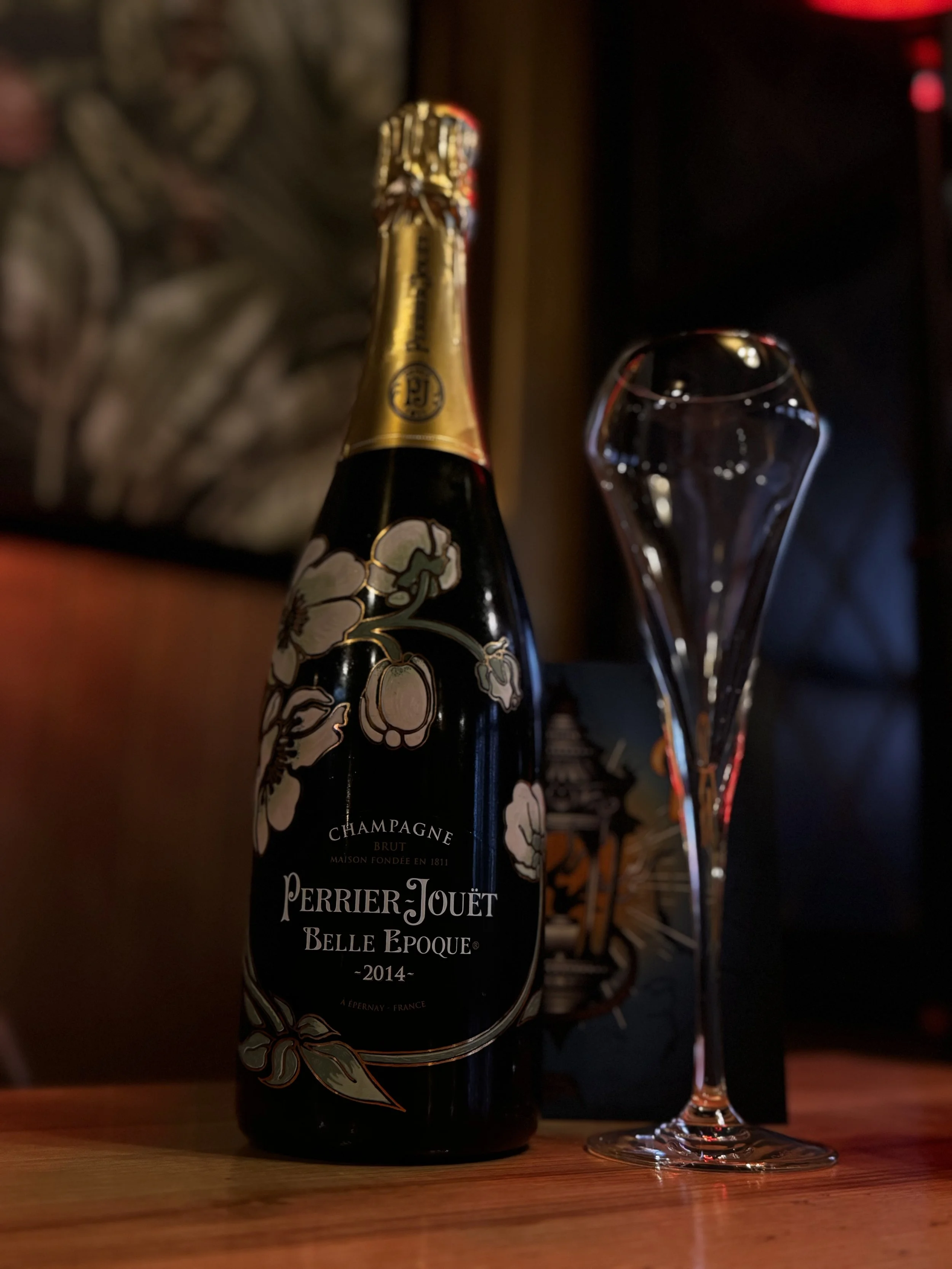 A bottle of Perrier-Jouët Belle Epoque champagne from 2014 with a floral label design, placed on a wooden surface next to an empty champagne flute. In the background, there is a dark gift box and a blurred decorative wall.