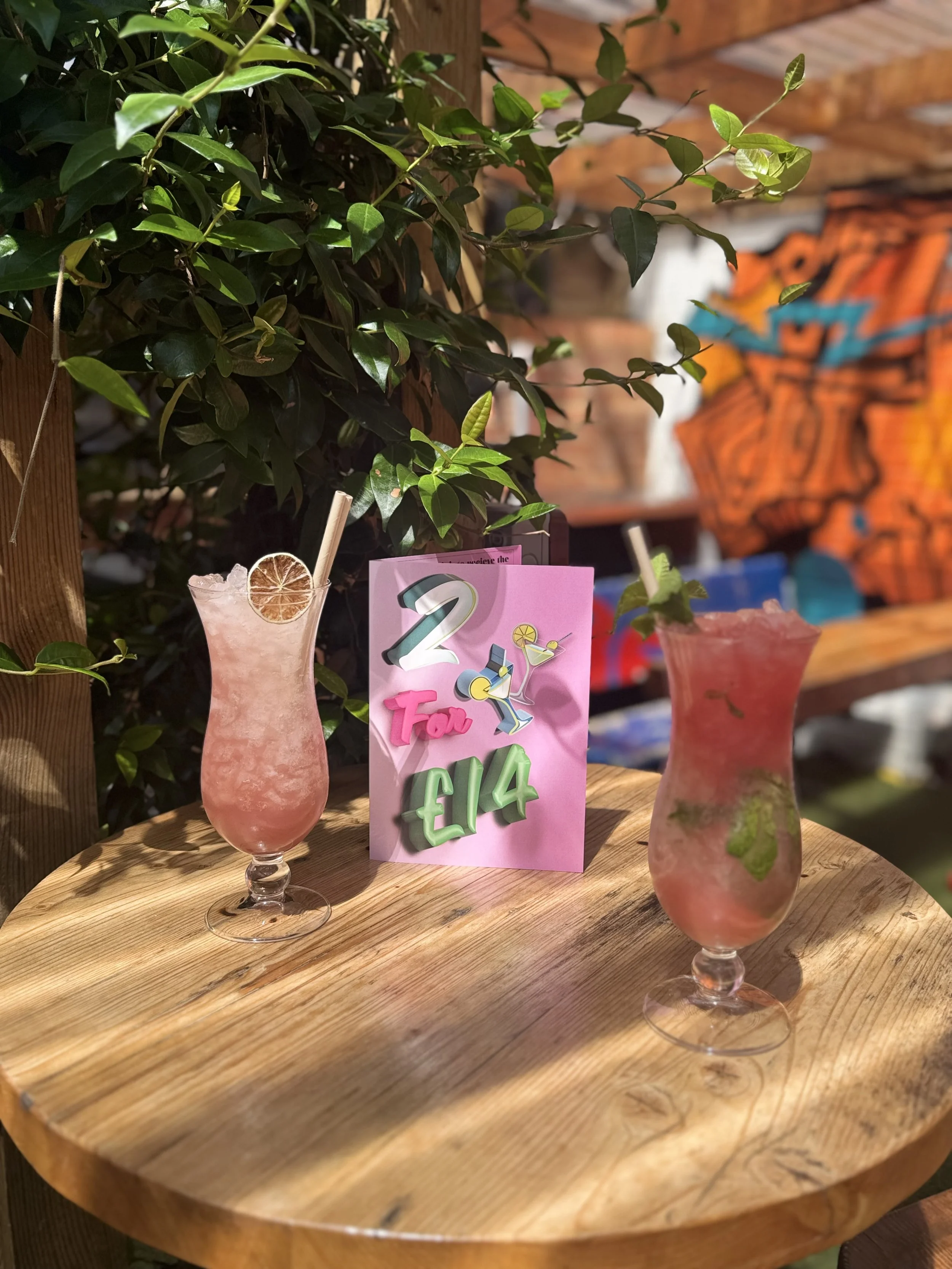 Two pink tropical cocktails with slices of lime or lemon and paper straws on a wooden table. A pink sign with colorful lettering and illustrations of drinks, indicating a special offer of 2 for €14. Background includes green foliage and a blurred wal
