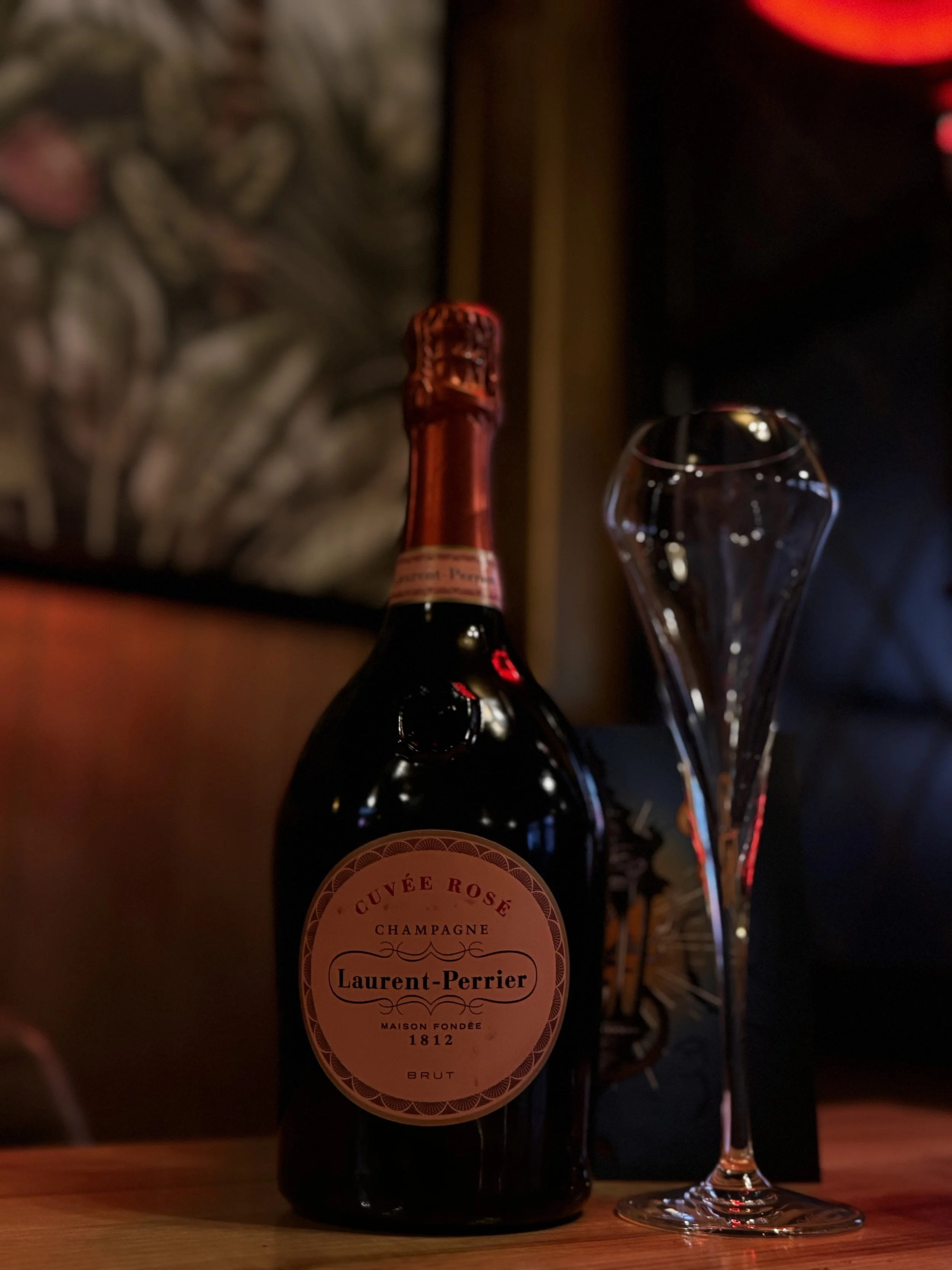 A bottle of Laurent-Perrier champagne and an empty flute glass on a wooden table in a dimly-lit room.