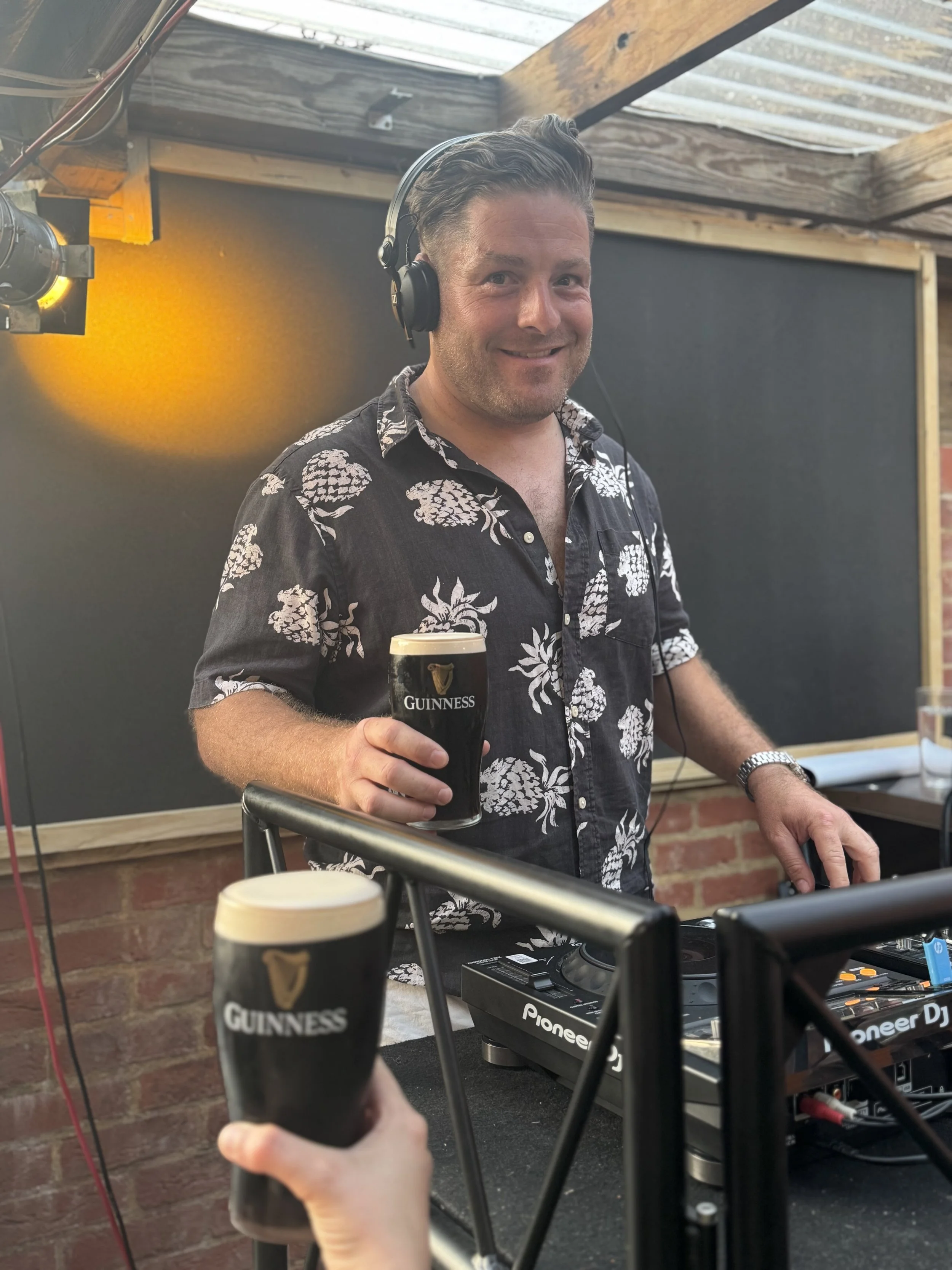 Man in a Hawaiian shirt smiling and holding a pint of Guinness beer, DJ equipment on the table, and another person holding a Guinness beer. The setting appears to be an outdoor or semi-outdoor space with a brick wall and a rustic ceiling.