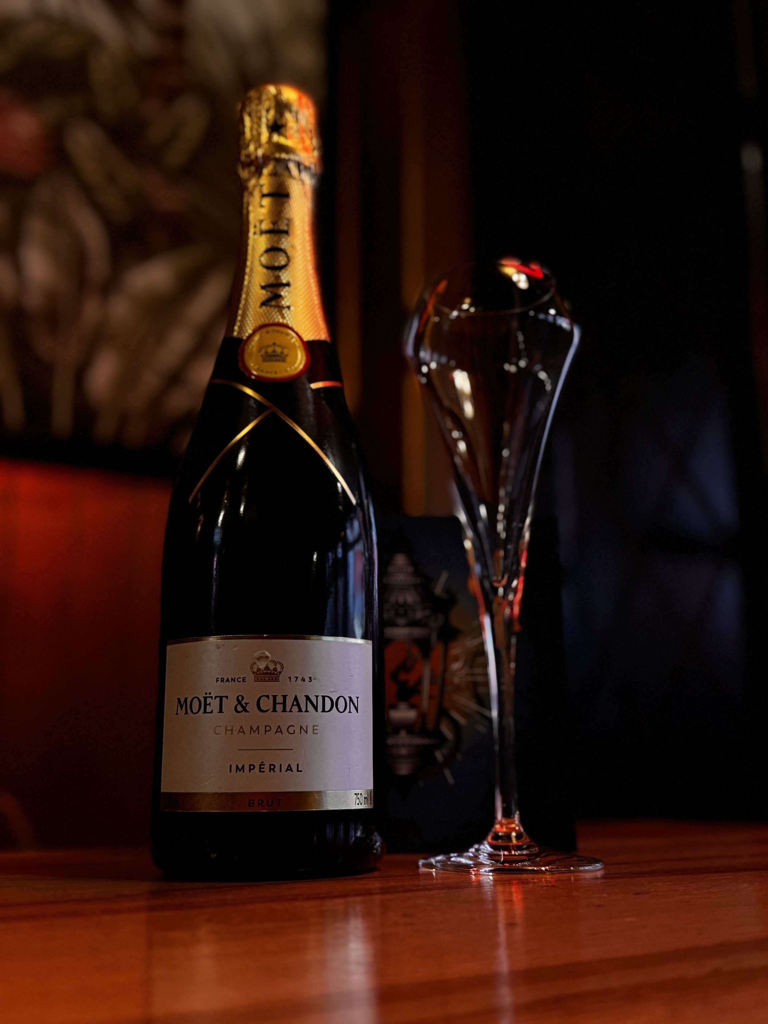A bottle of Moët & Chandon Champagne Imperial Brunt with a tall, empty champagne flute on a wooden surface in a dimly lit setting.