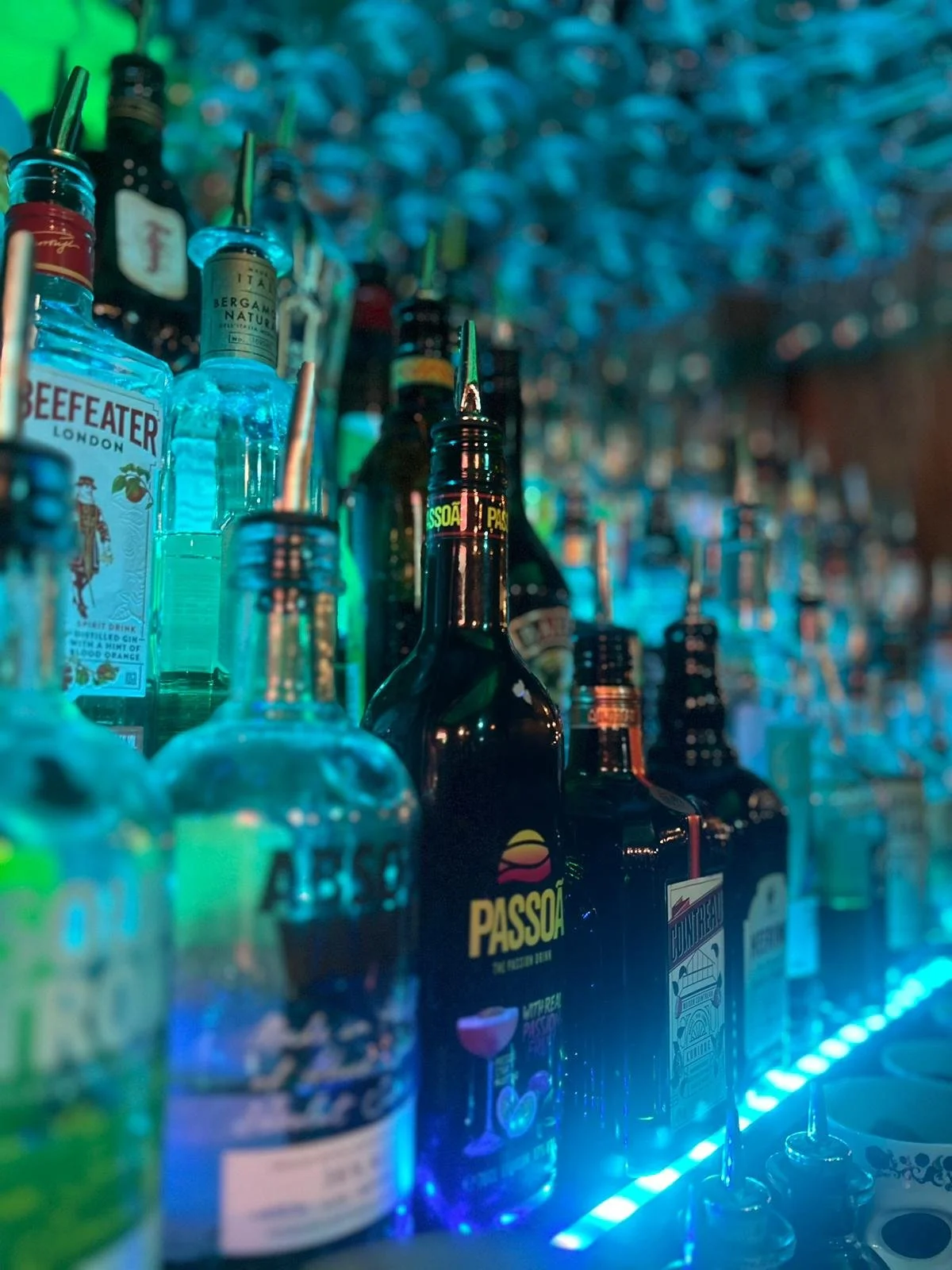 A row of various liquor bottles on a bar shelf illuminated by colorful lights, including brands like Beefeater, Passoa, and others, with a blurred background.