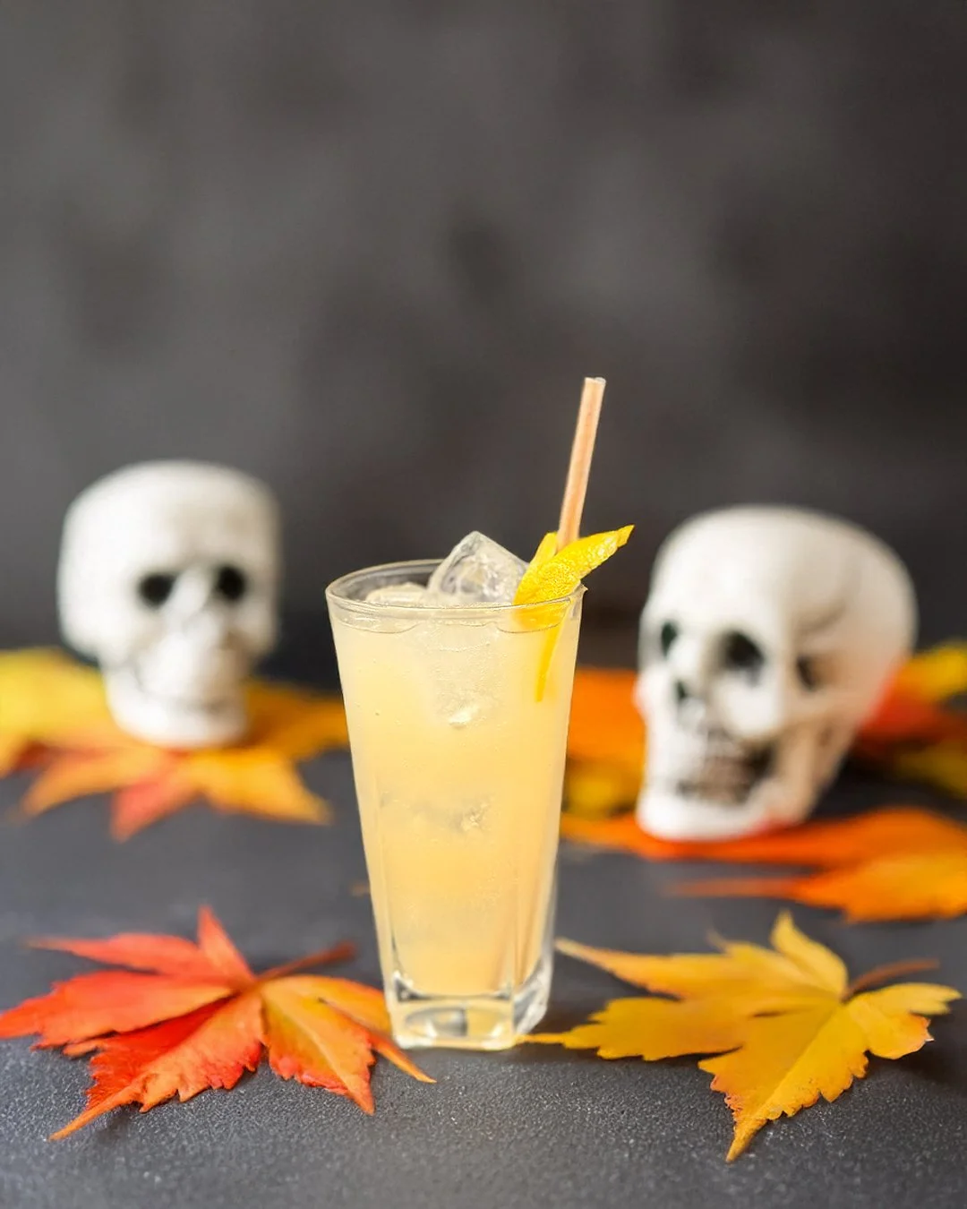 Yellow fruity cocktail with ice and lemon slice garnished, surrounded by colourful autumn leaves, with two white skull props in the background on a dark surface. Halloween cocktails.