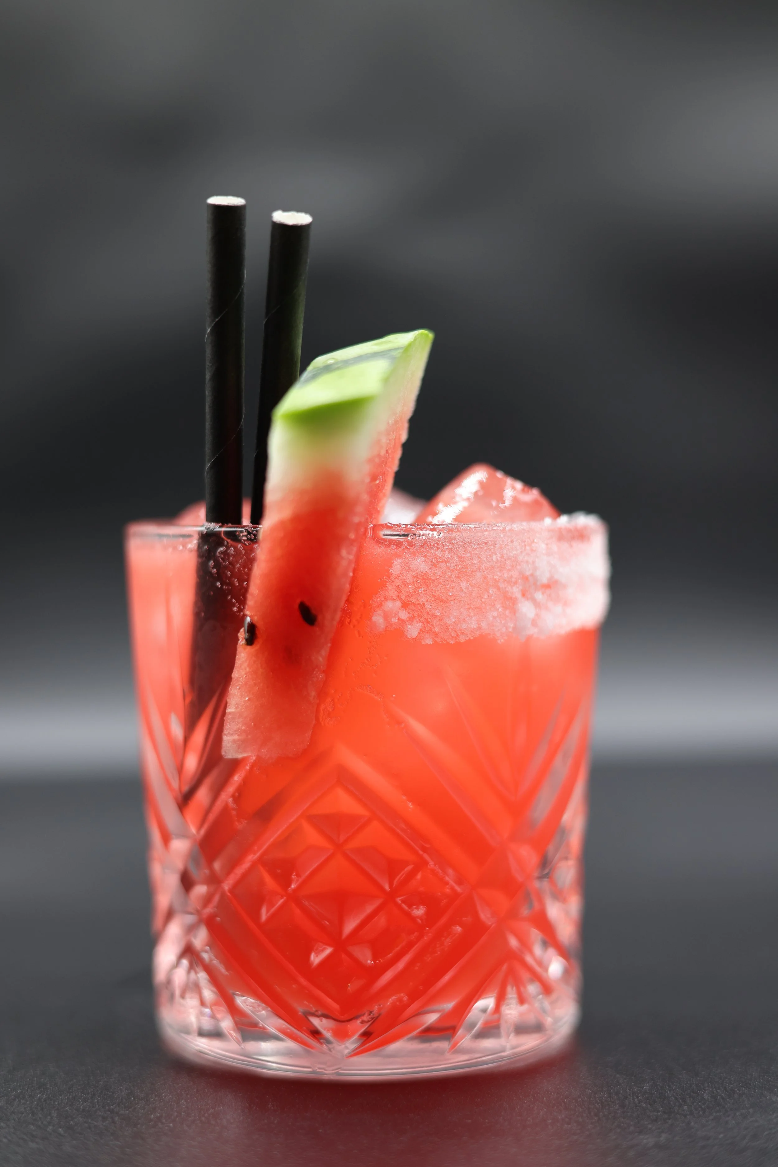 A crystal glass filled with a pink cocktail, garnished with a slice of watermelon and a slice of cucumber, with two black straws.