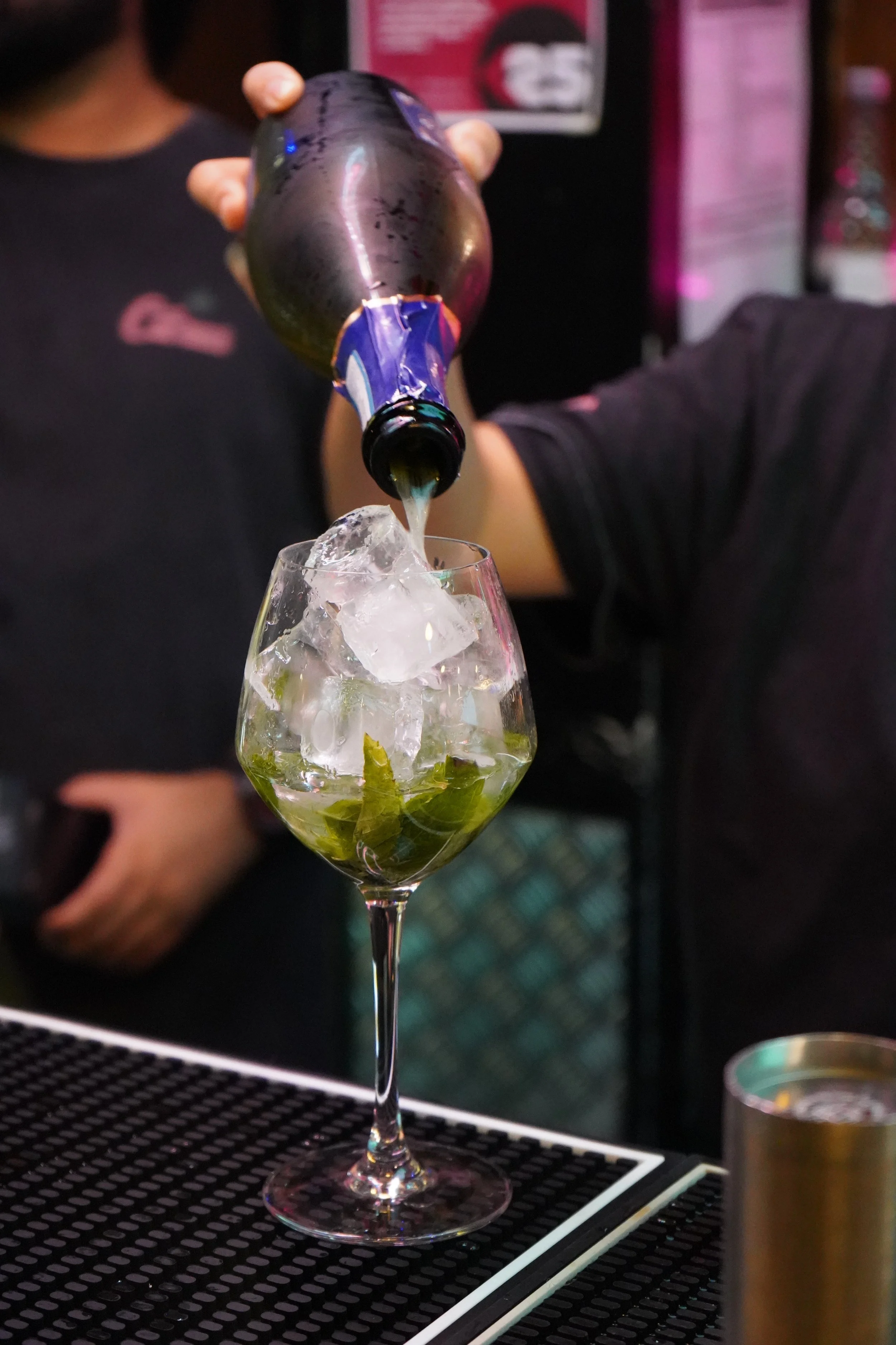 Bartender pouring a dark bottle of beverage into a glass filled with ice and mint leaves.
