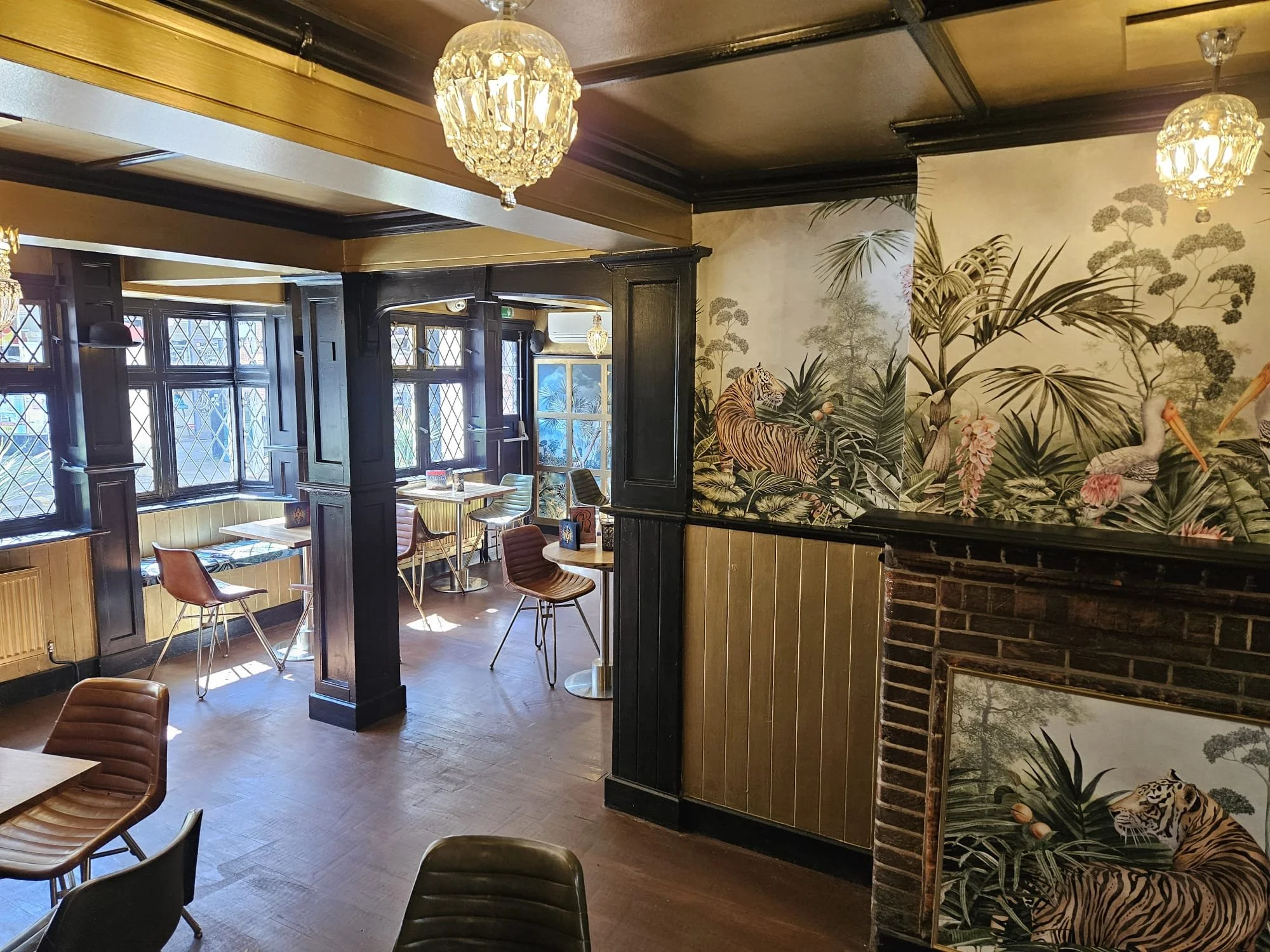 Interior of a cafe or restaurant with wooden floors, wooden paneling, a mural with jungle animals and plants, large windows with diamond-patterned panes, and various chairs and tables.
