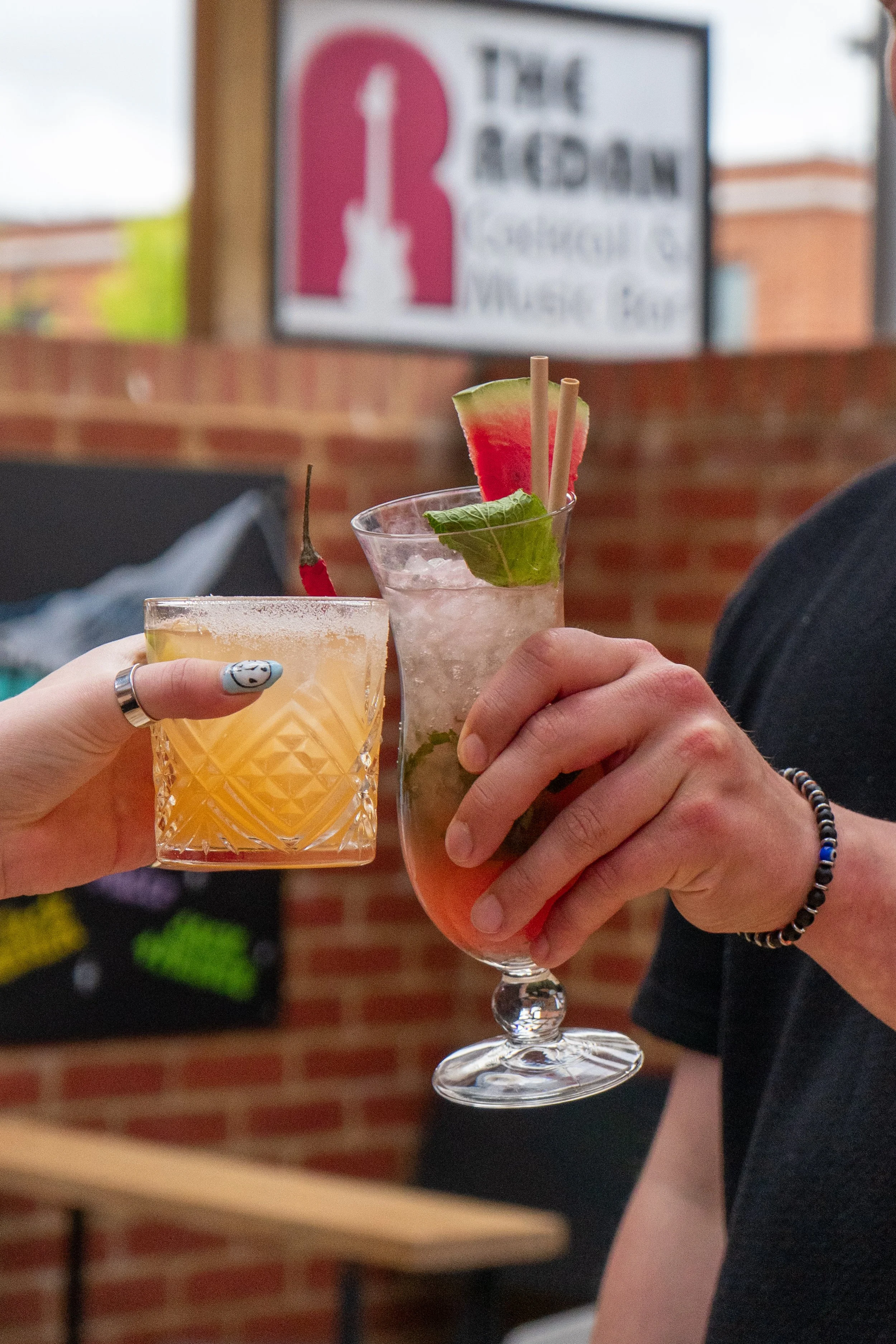 Two people clinking cocktails, a short glass with a yellow drink and a tall glass with a pink drink garnished with watermelon, lime, and basil, in a casual indoor setting with brick walls.