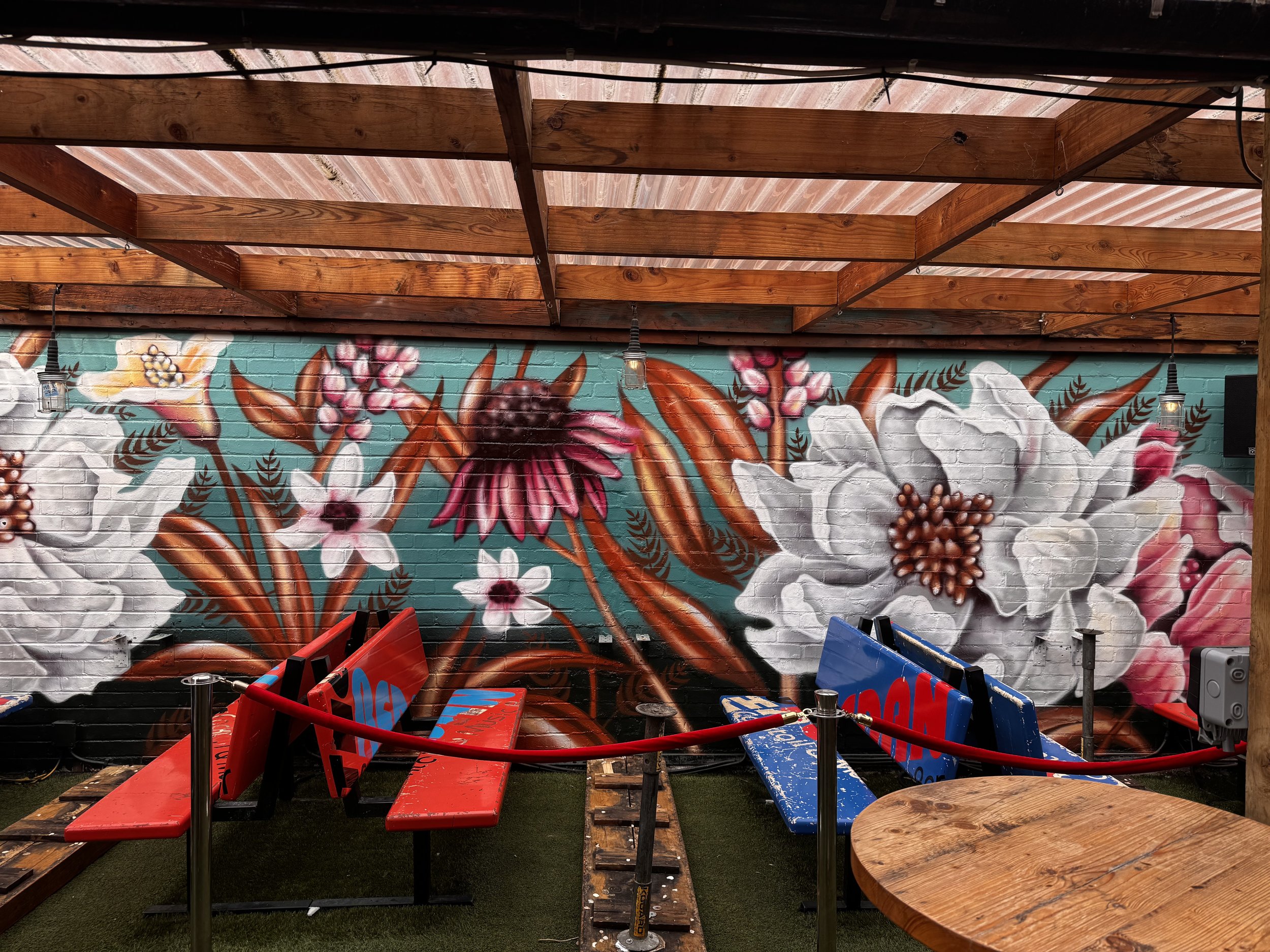 Colorful mural of flowers and leaves on a brick wall in a patio area with red and blue benches and wooden tables.