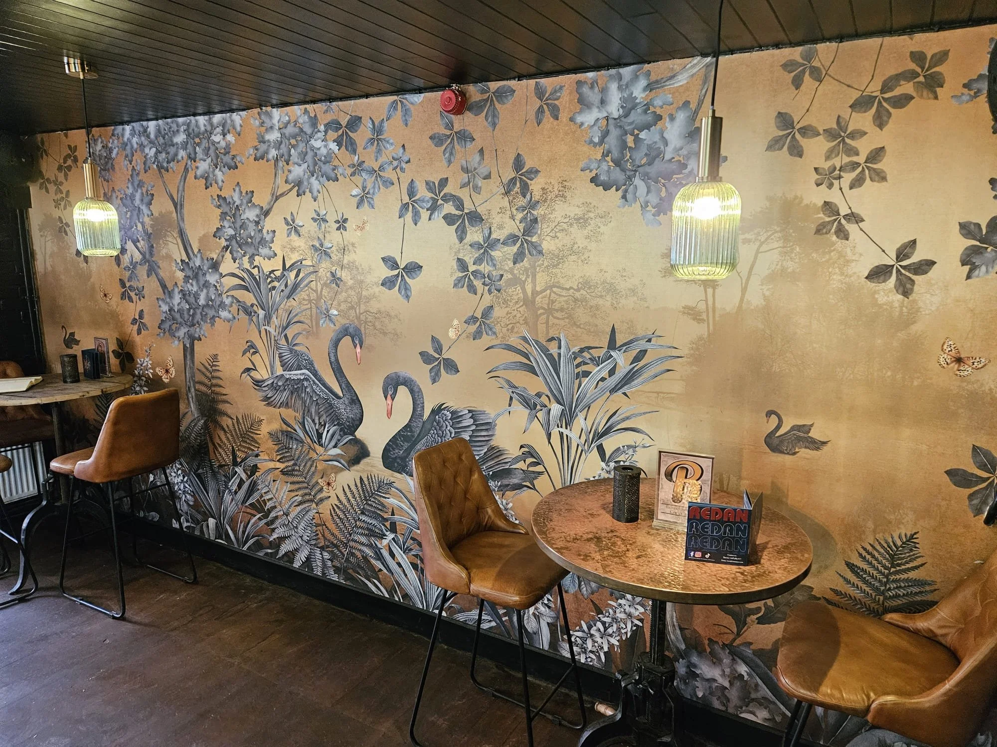 Interior of a restaurant or cafe with a detailed mural of swans, trees, and foliage on the wall, illuminated by hanging pendant lights, with tables and chairs arranged for seating.