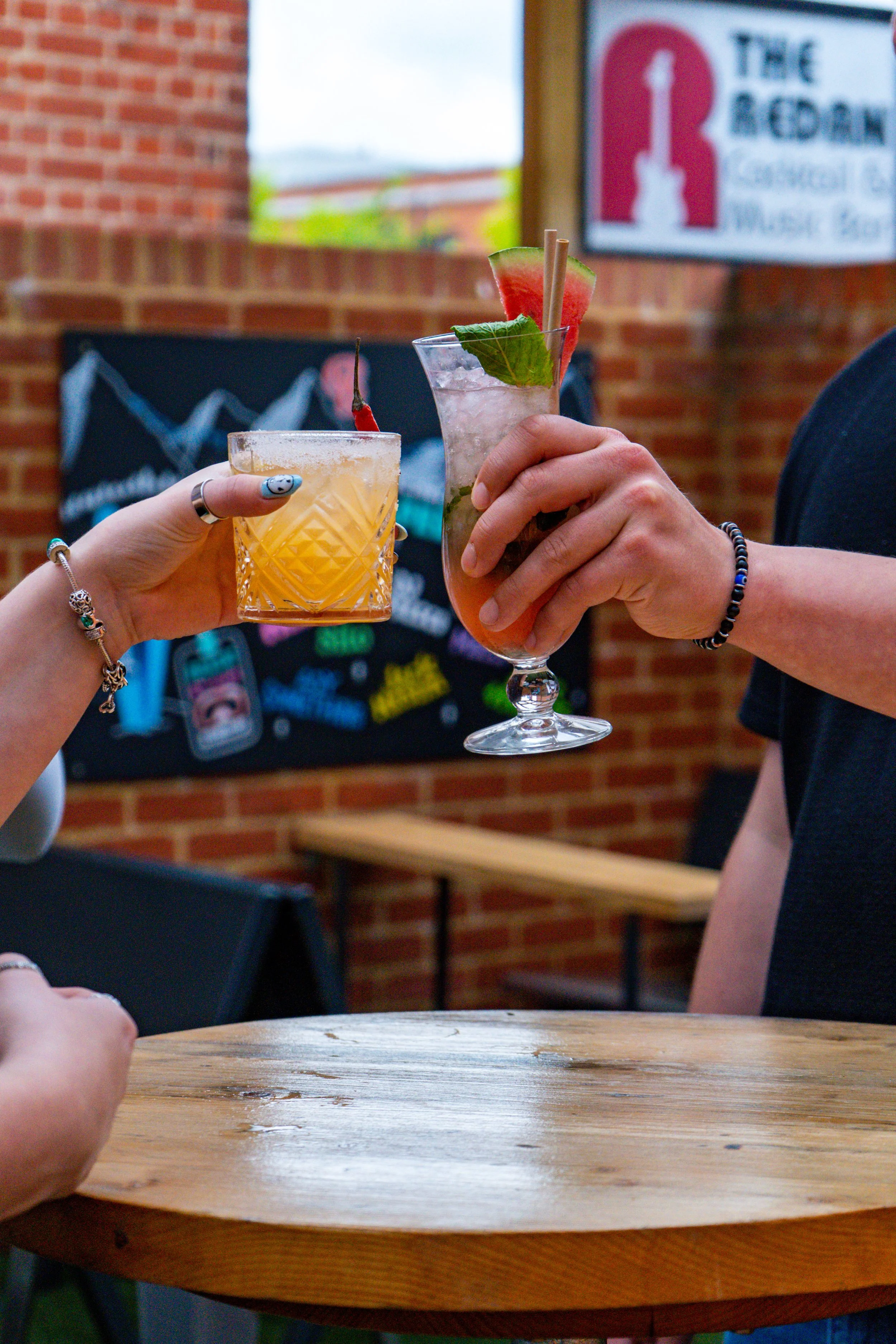 Two people clinking colorful cocktails, one with a watermelon garnish and the other with a cherry, at a bar or restaurant.