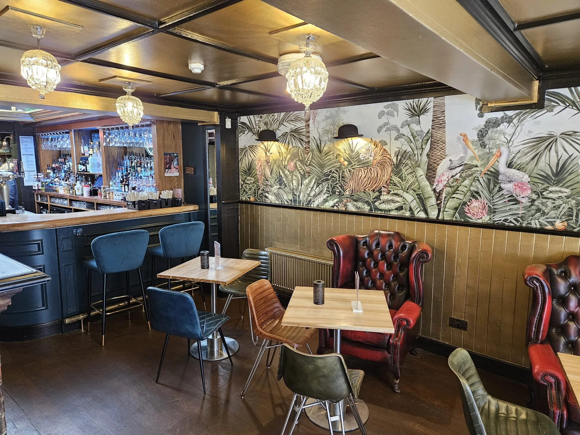 Interior of a bar and dining area with vintage leather chairs, small tables, a wallpaper mural of a jungle scene with tigers and flamingos, a wooden bar counter, and hanging chandeliers.