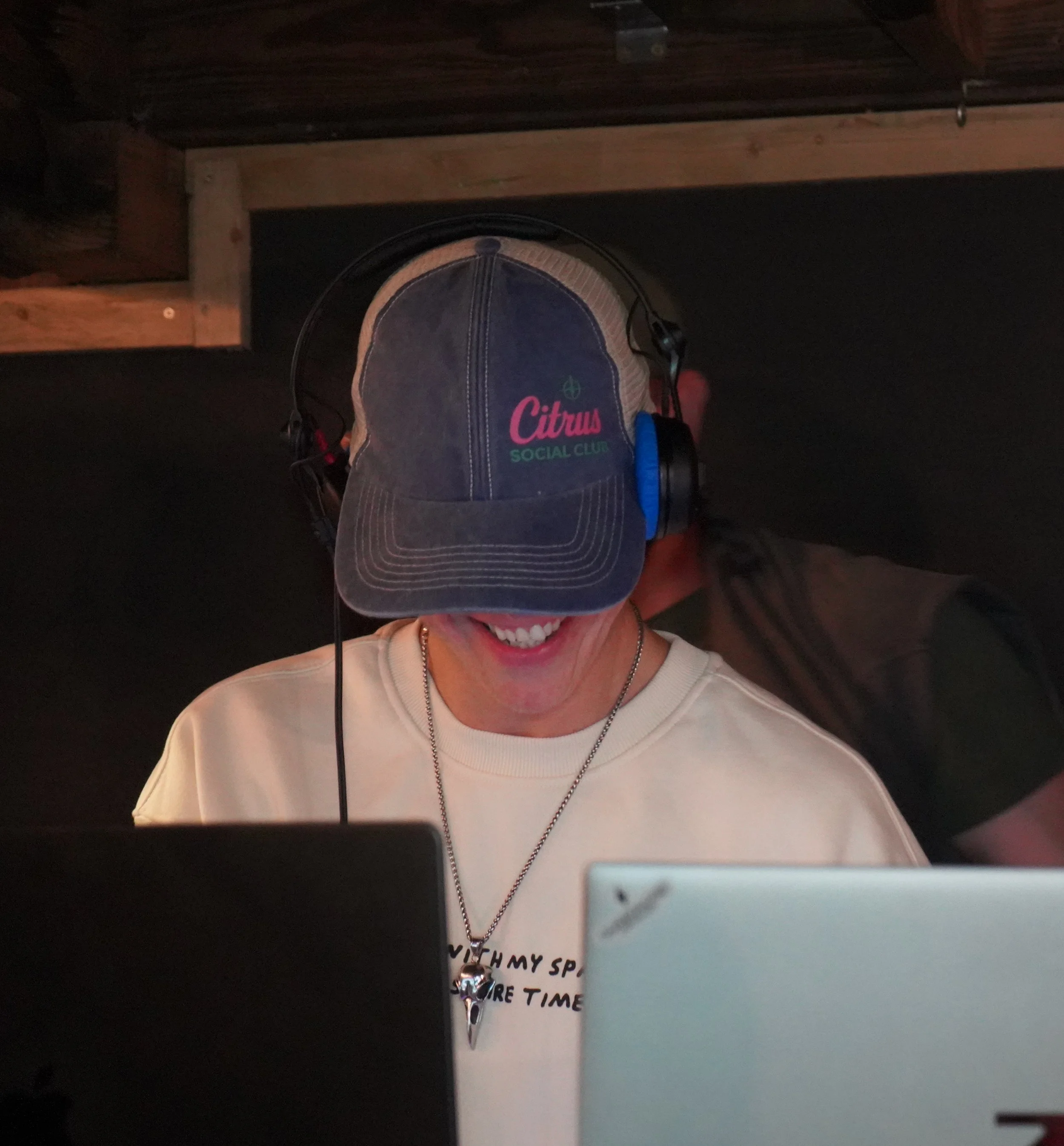 a DJ wearing a 'Citrus Social Club' T-shirt brand branded cap headphones, smiling, in front of a computer and DJ decks.