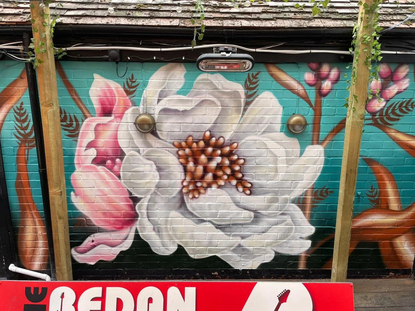 A large mural of a white flower with pink and brown accents painted on a brick wall, with some greenery on the sides and outdoor lights attached to the wall.