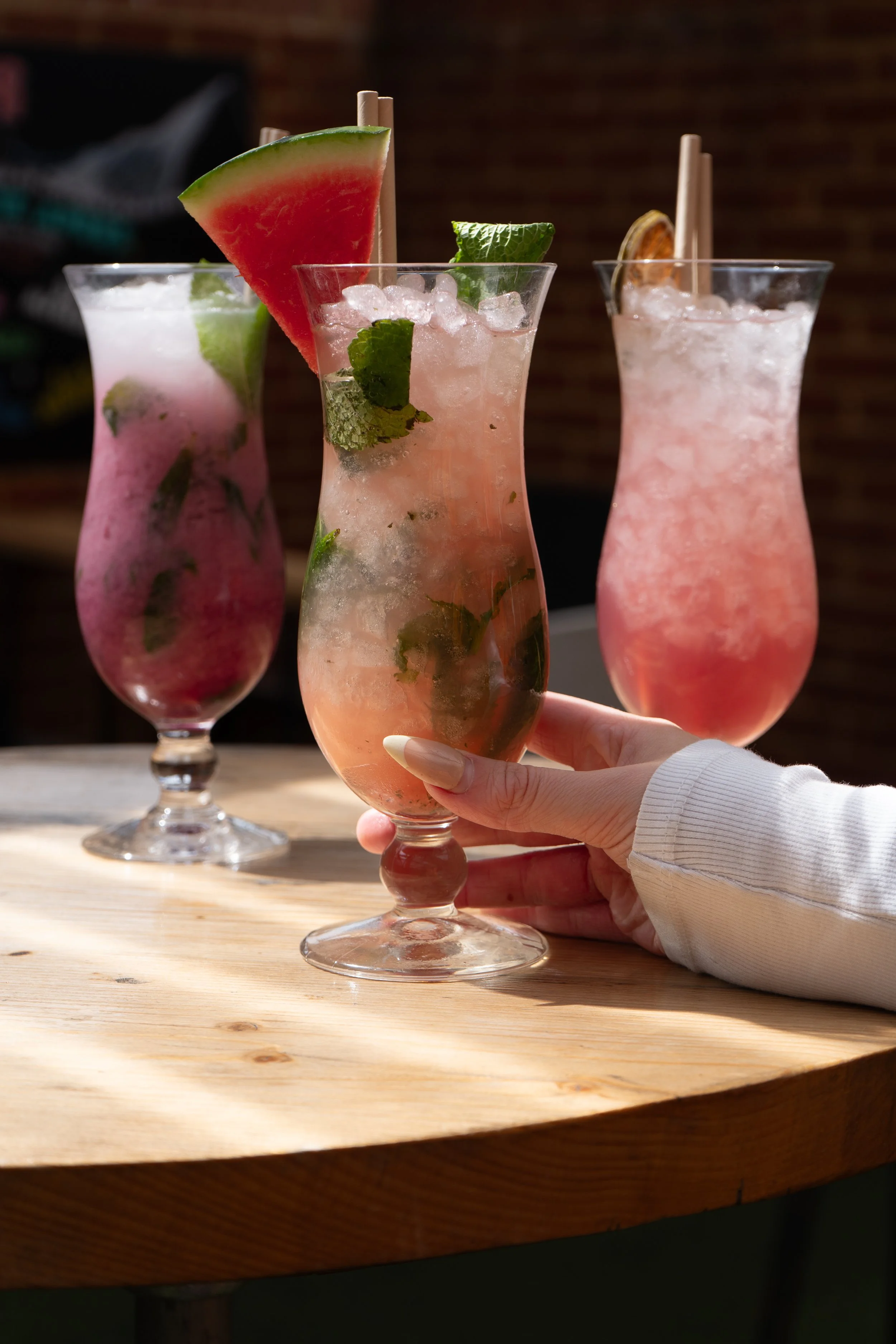 Three colorful cocktails with fruit garnishes on a wooden table, one person holding the front glass.