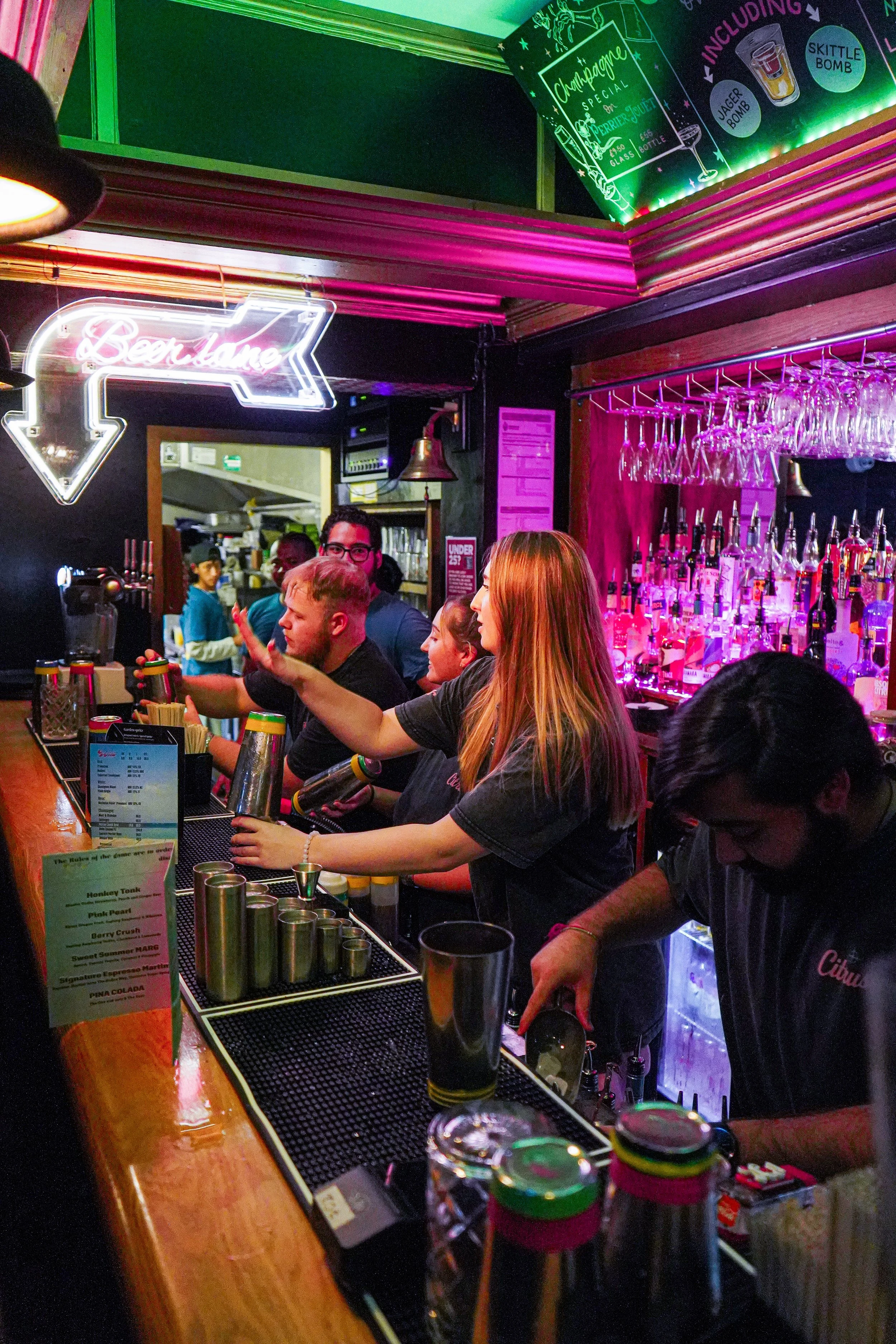 People working behind a nightclub bar with colorful neon signs, bar tools, and liquor bottles, serving drinks to customers.