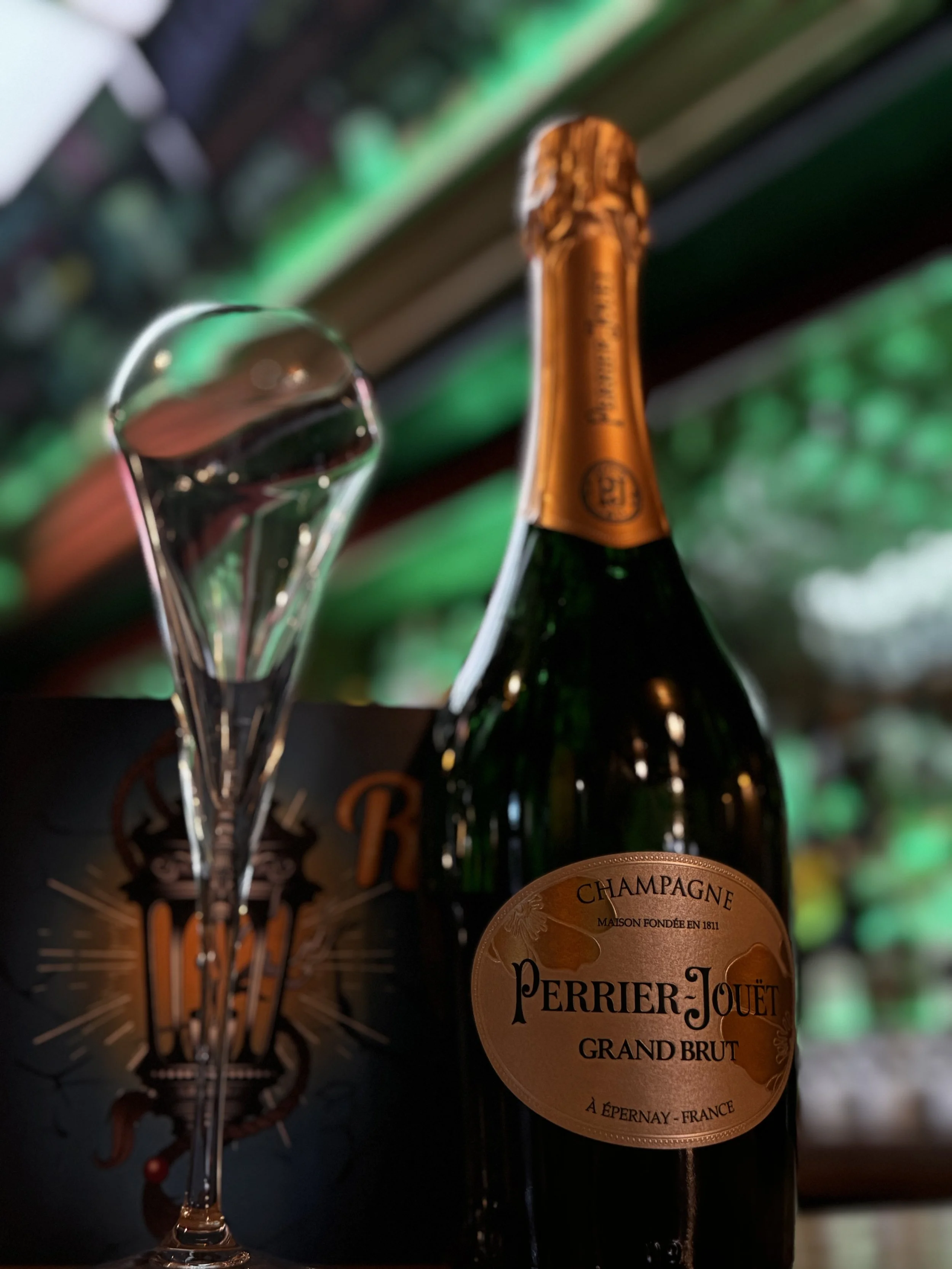 A bottle of Perrier-Jouët Grand Brut champagne with a champagne flute next to it, set against a background with green and warm lighting.