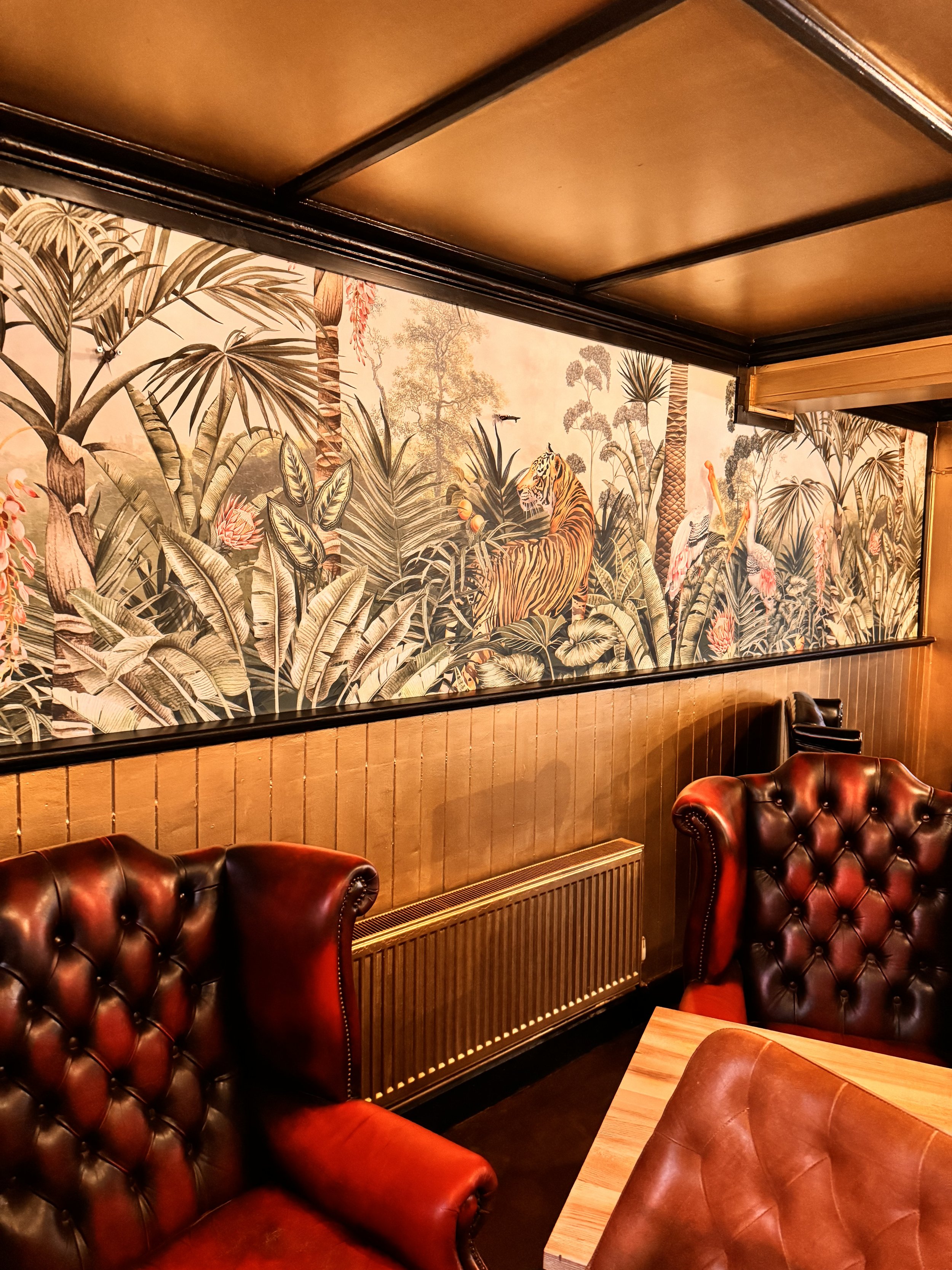 Interior of a restaurant with tropical-themed wallpaper featuring a tiger, flamingos, and lush foliage. There are large, brown leather armchairs with tufted backs, a wooden table, and wood-paneled walls and ceiling.