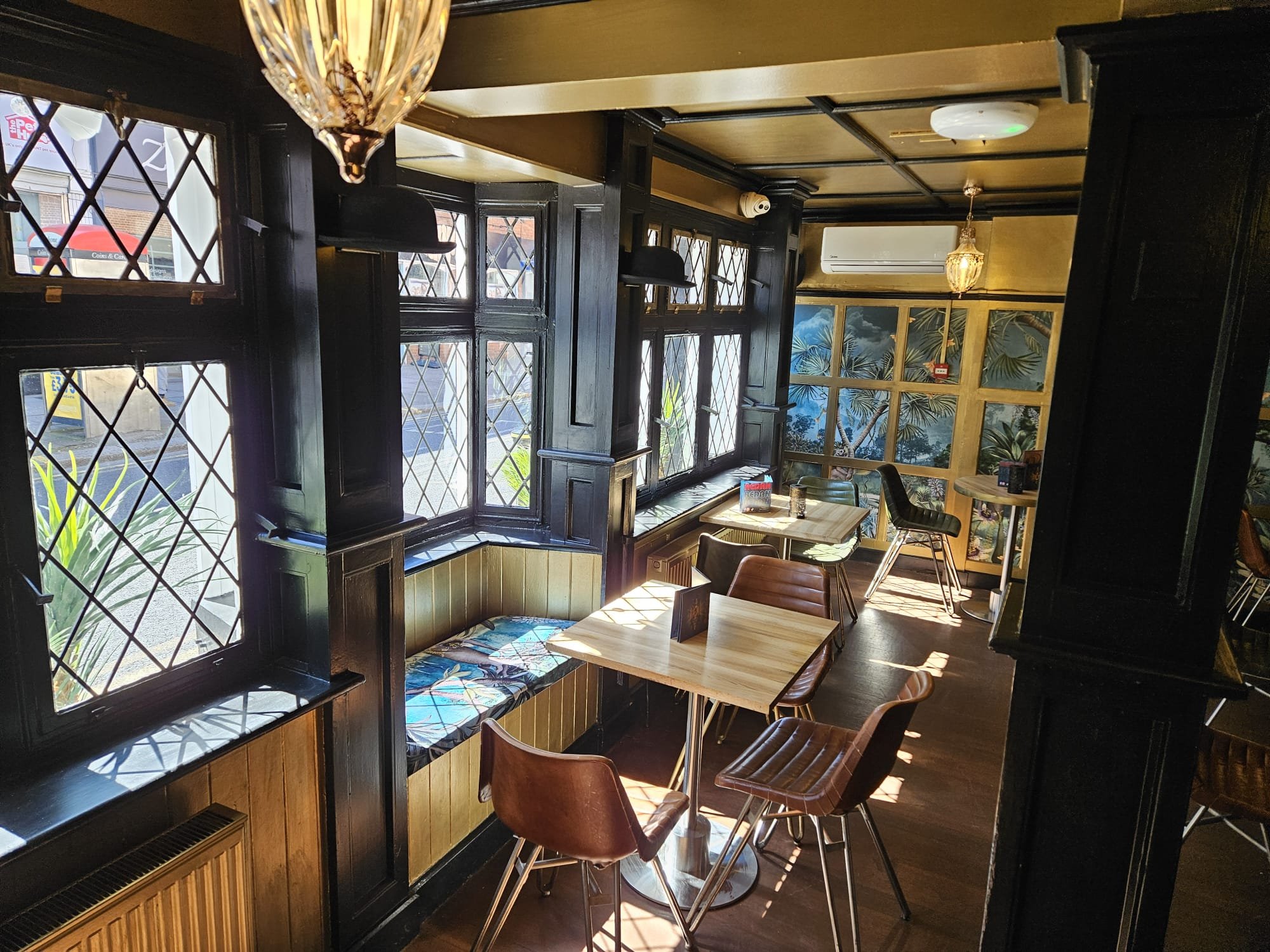 Sunny interior of a cozy café with wooden tables and brown chairs, black window frames with diamond-shaped panes, tropical-themed wall murals, and decorative pendant lighting.