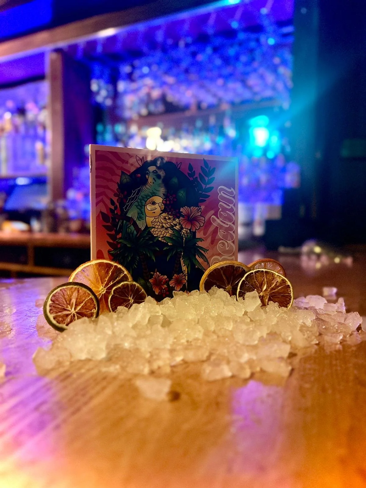 A colorful cocktail menu card placed in the center of a pile of crushed ice on a bar counter, surrounded by lemon and lime slices, with a blurred bar background illuminated by blue and purple neon lights.