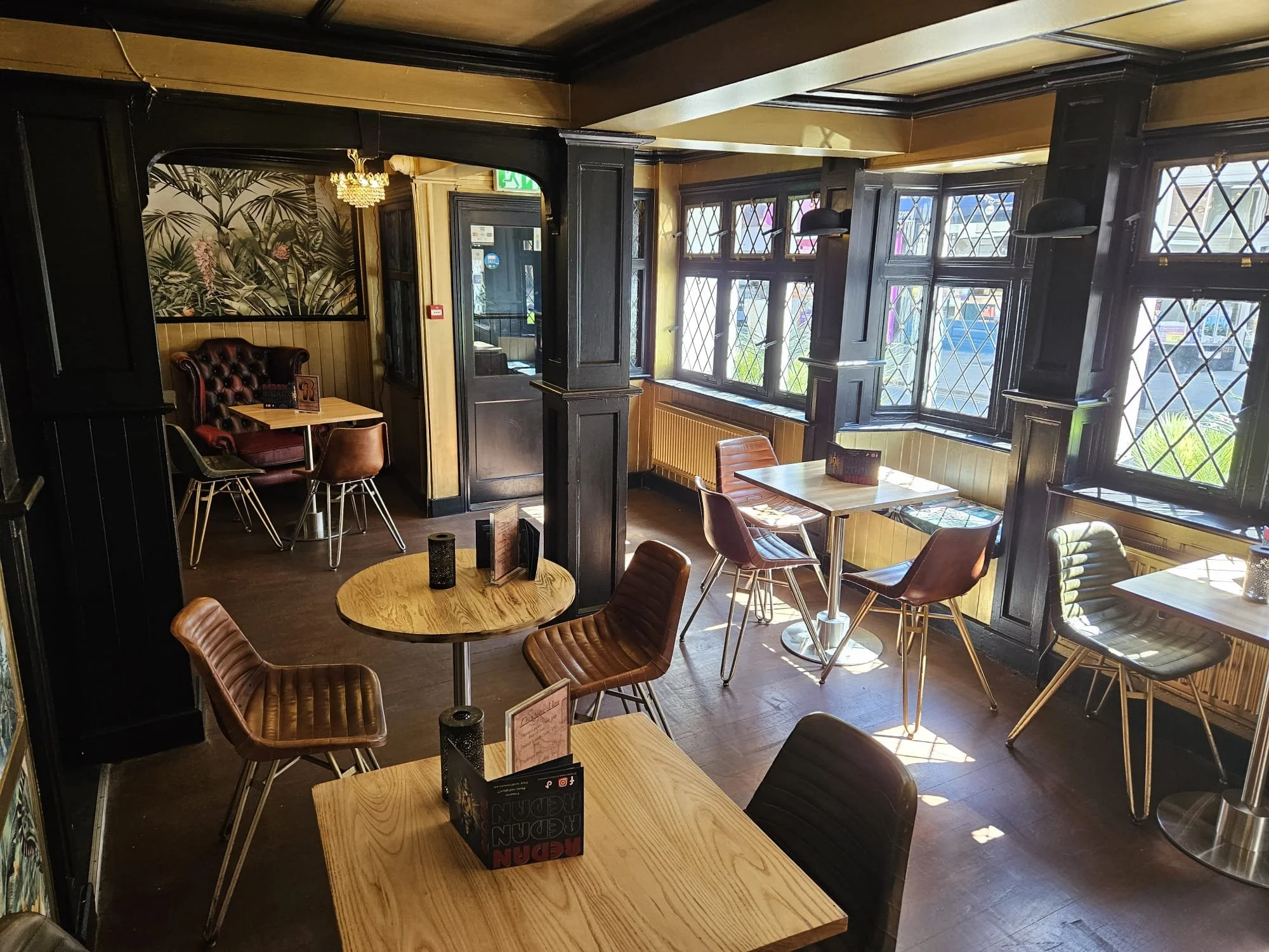 Interior of a cozy cafe with wooden tables and leather chairs, black wood paneling, large windows with diamond-shaped panes, and a mural with botanical design on the wall.