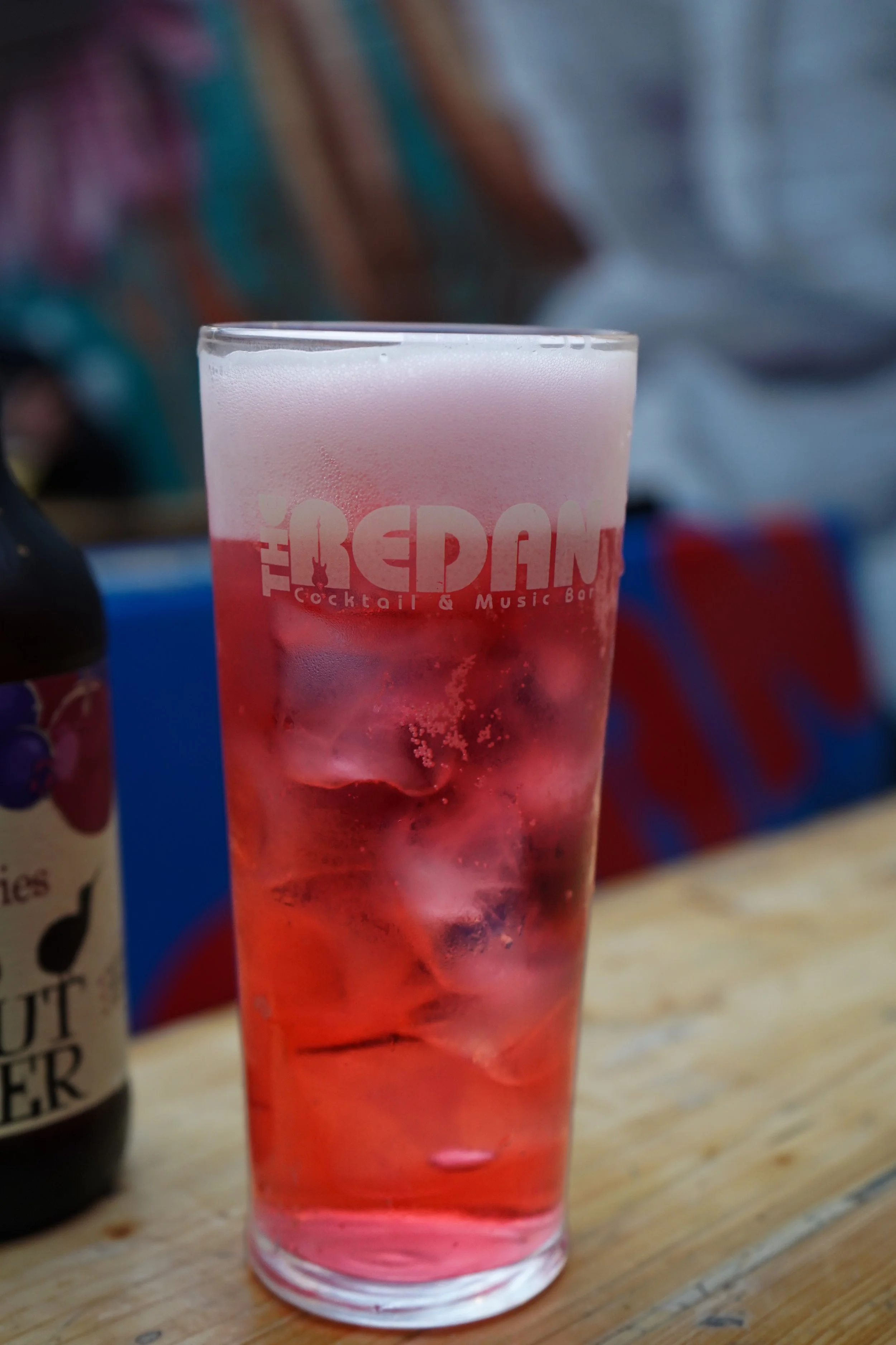 A tall glass with a pink beverage and ice cubes, labeled 'The Bedlam' cocktail and music bar, placed on a wooden table.