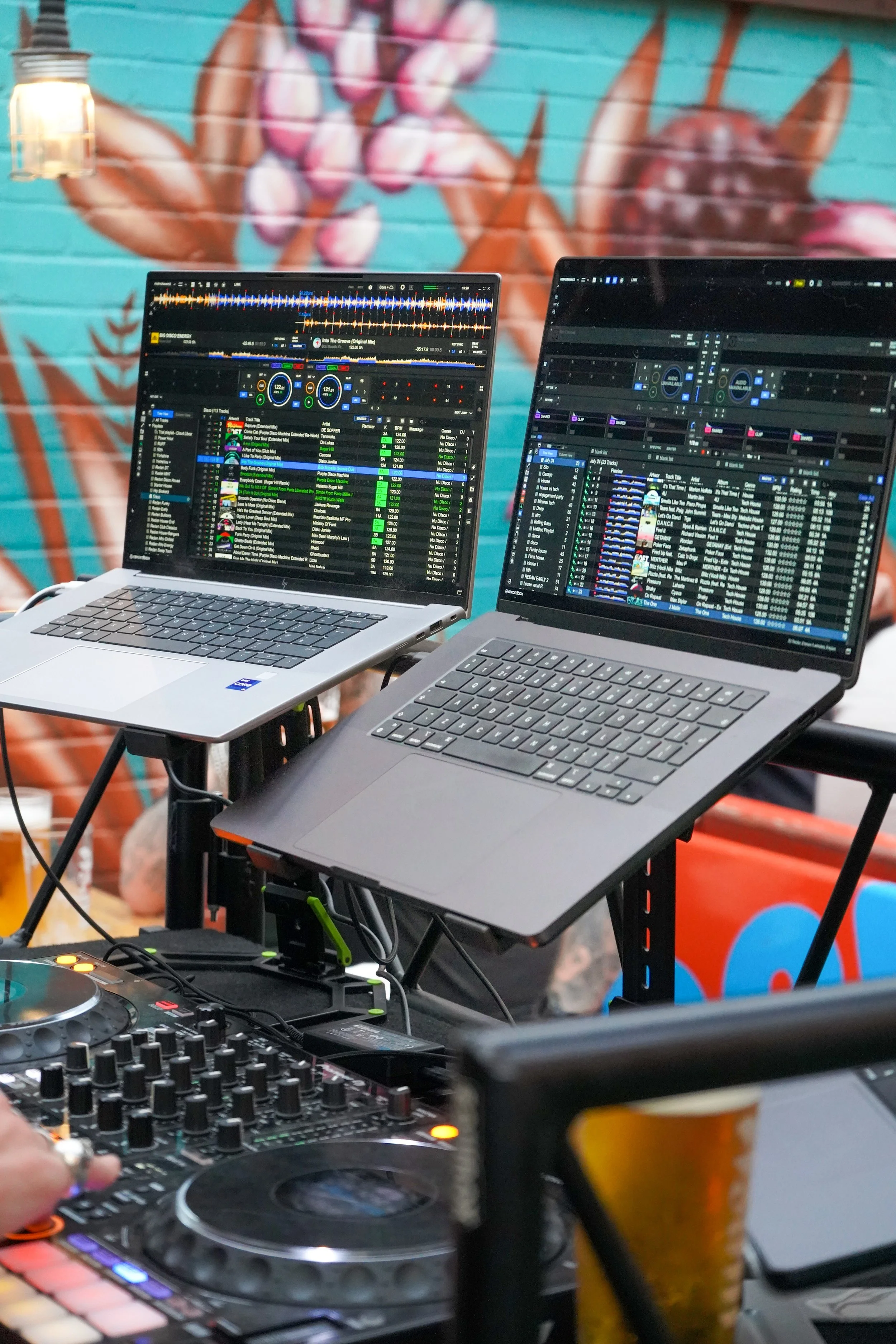 Two laptop computers with DJ software running on their screens, placed on a DJ booth with a mixer and controller, set against a colorful mural background.