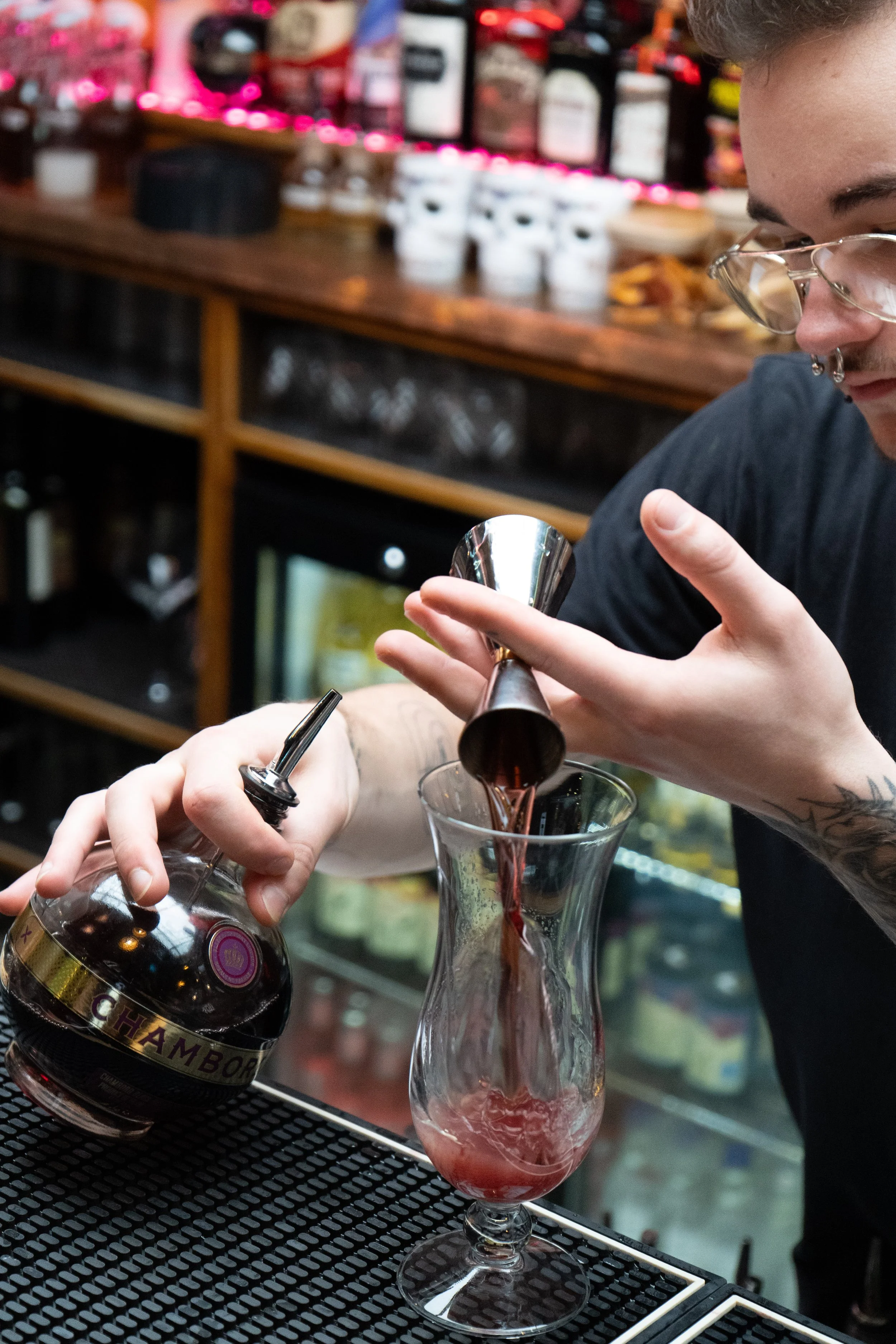 A bartender preparing a layered cocktail with a jigger and a bitters bottle in a bar setting.