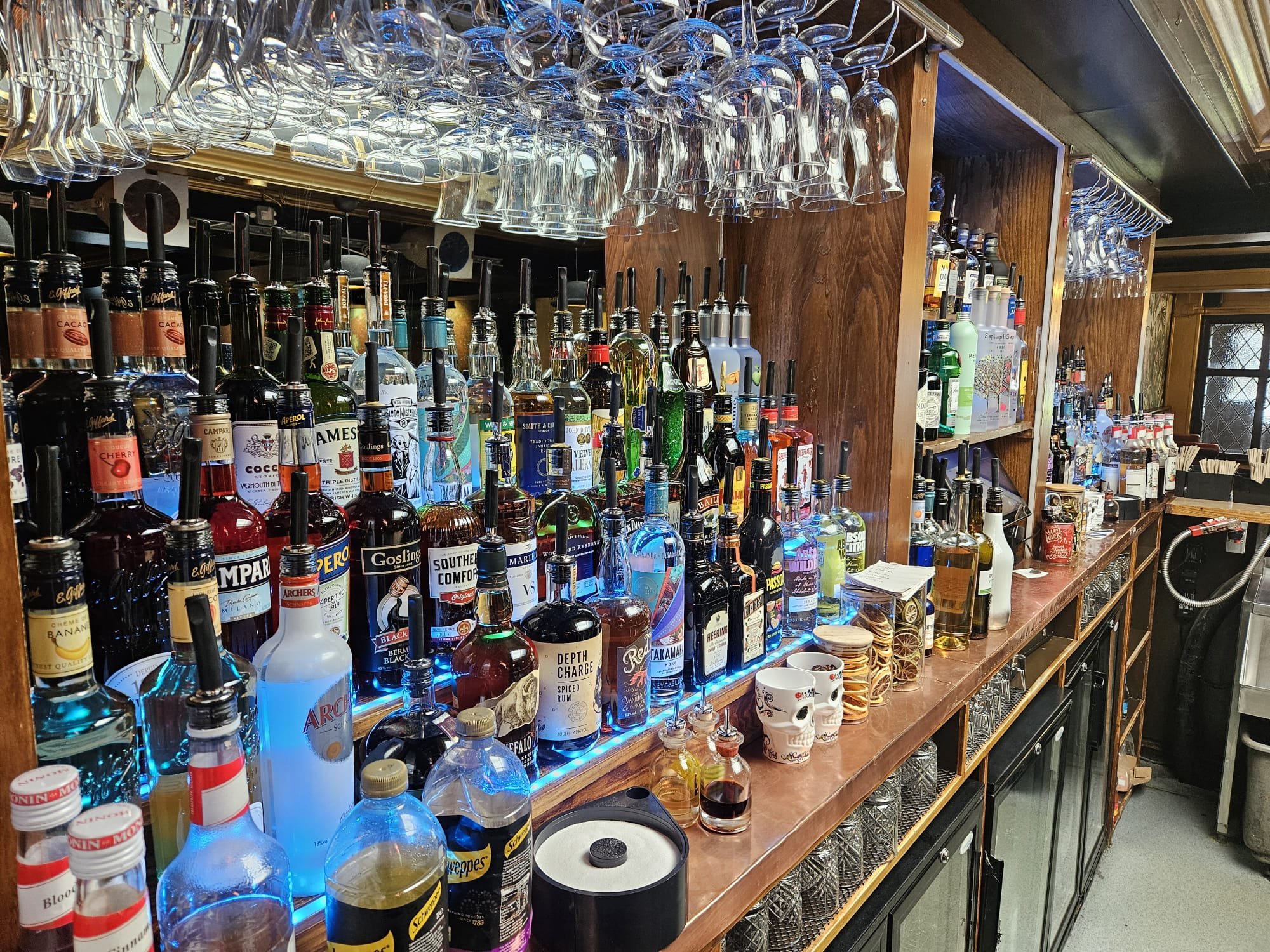 A bar counter with a wide assortment of liquor bottles, wine glasses hanging upside down above, and some decorative skull mugs on the counter.