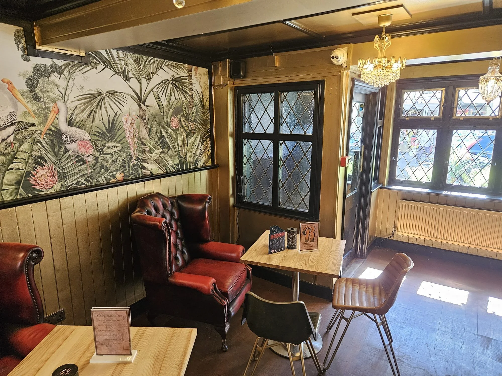 Interior of a cozy restaurant or cafe with red leather armchairs, wooden tables, and vintage-style windows with diamond-shaped panes. There is a large botanical wallpaper featuring birds and lush foliage on one wall, and a chandelier hanging from the