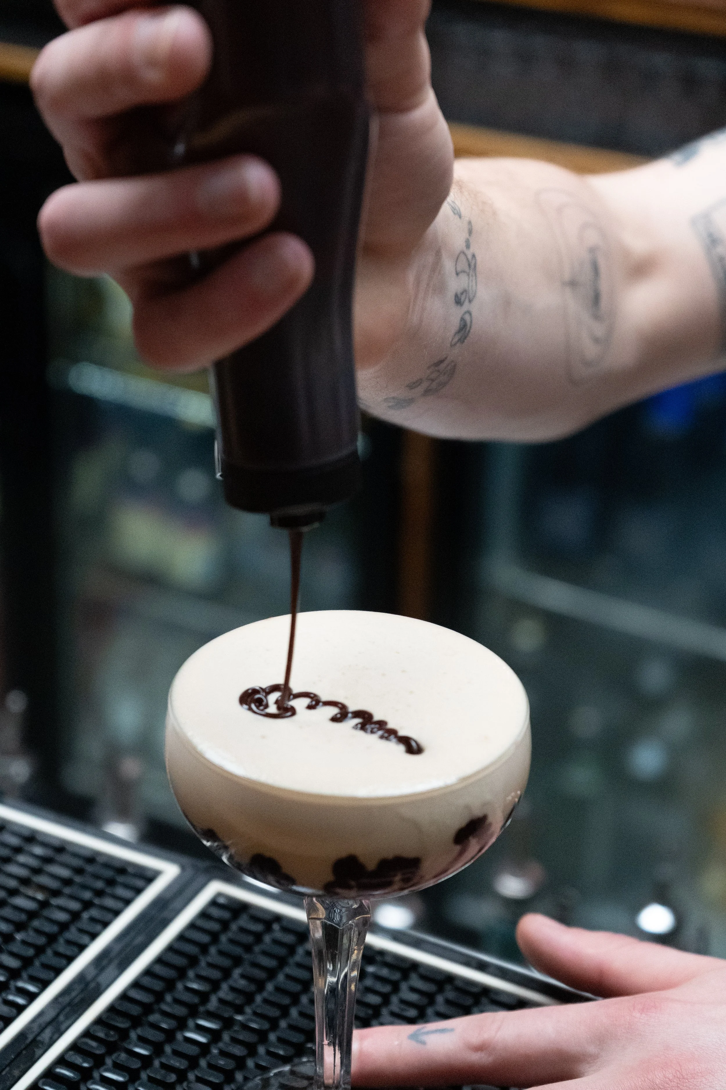 Bartender pouring chocolate syrup onto a white cocktail with black tapioca pearls, using a squeeze bottle.