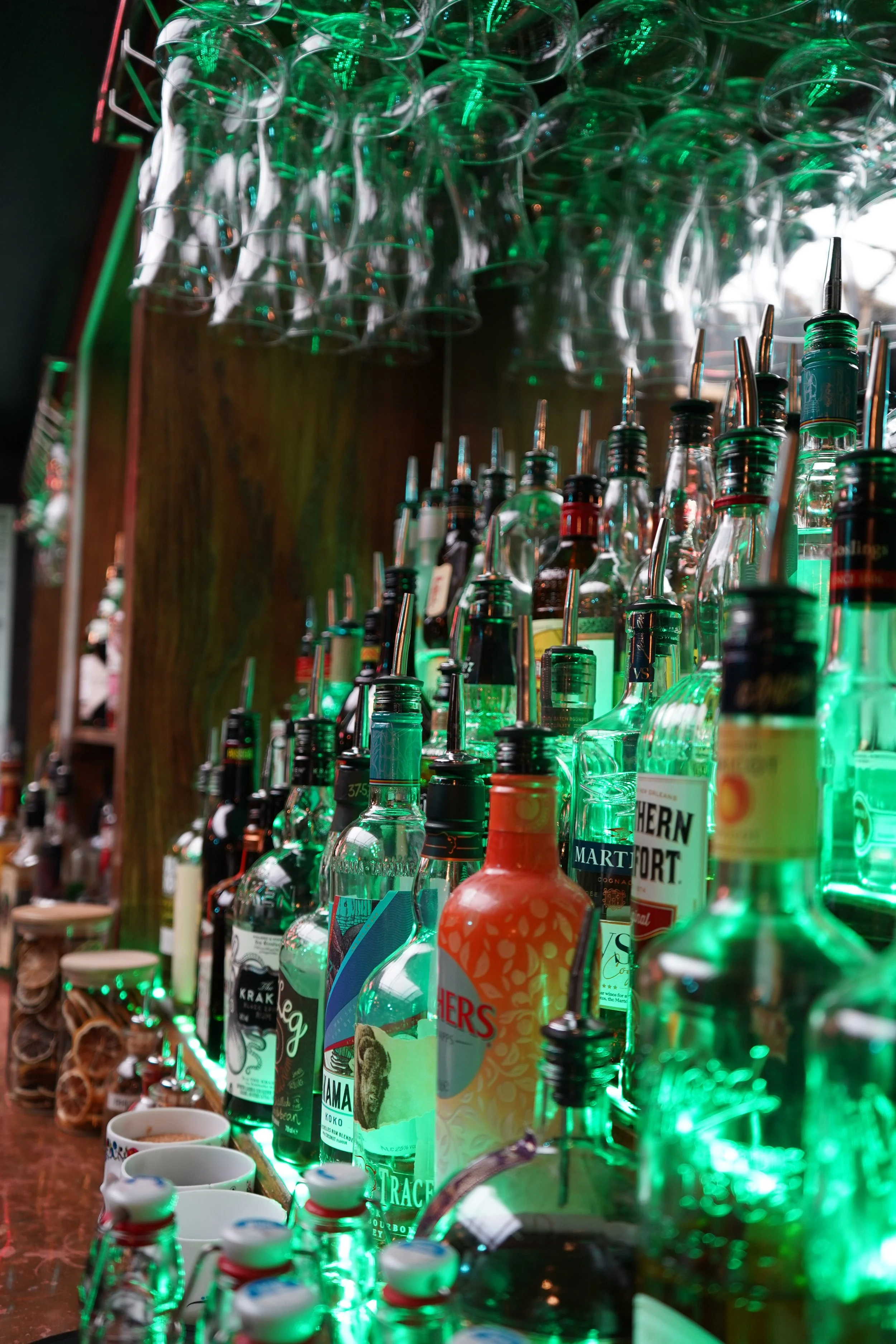 A bar shelf stocked with various liquors illuminated by green lights, cups, and bar accessories.