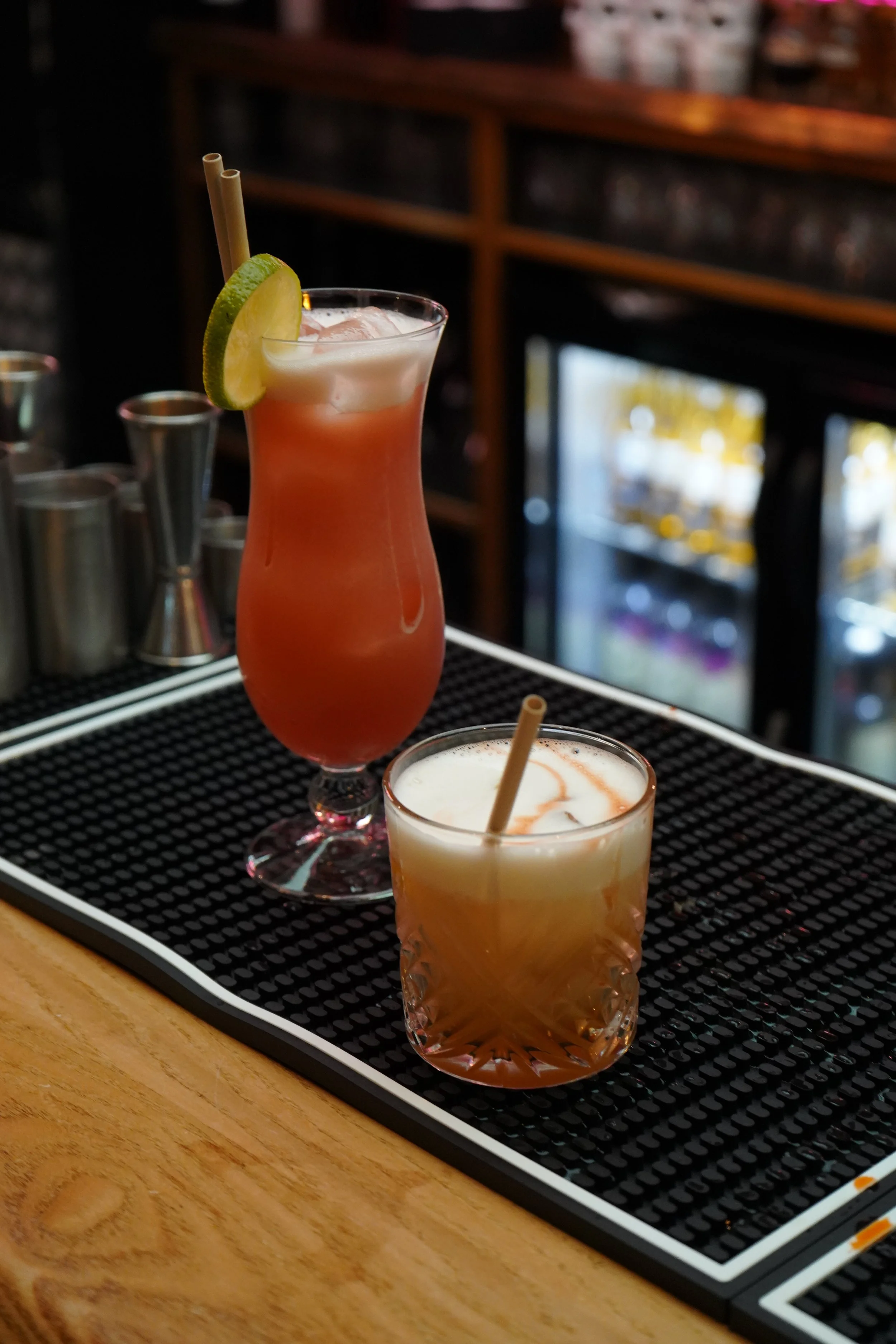 Two colorful cocktails on a black bar mat, one tall pinkish drink in a hurricane glass with a lime wedge and two straws, and a shorter Old Fashioned glass with yellowish drink with a straw, garnish involves vegan foamer, perfect foam. 