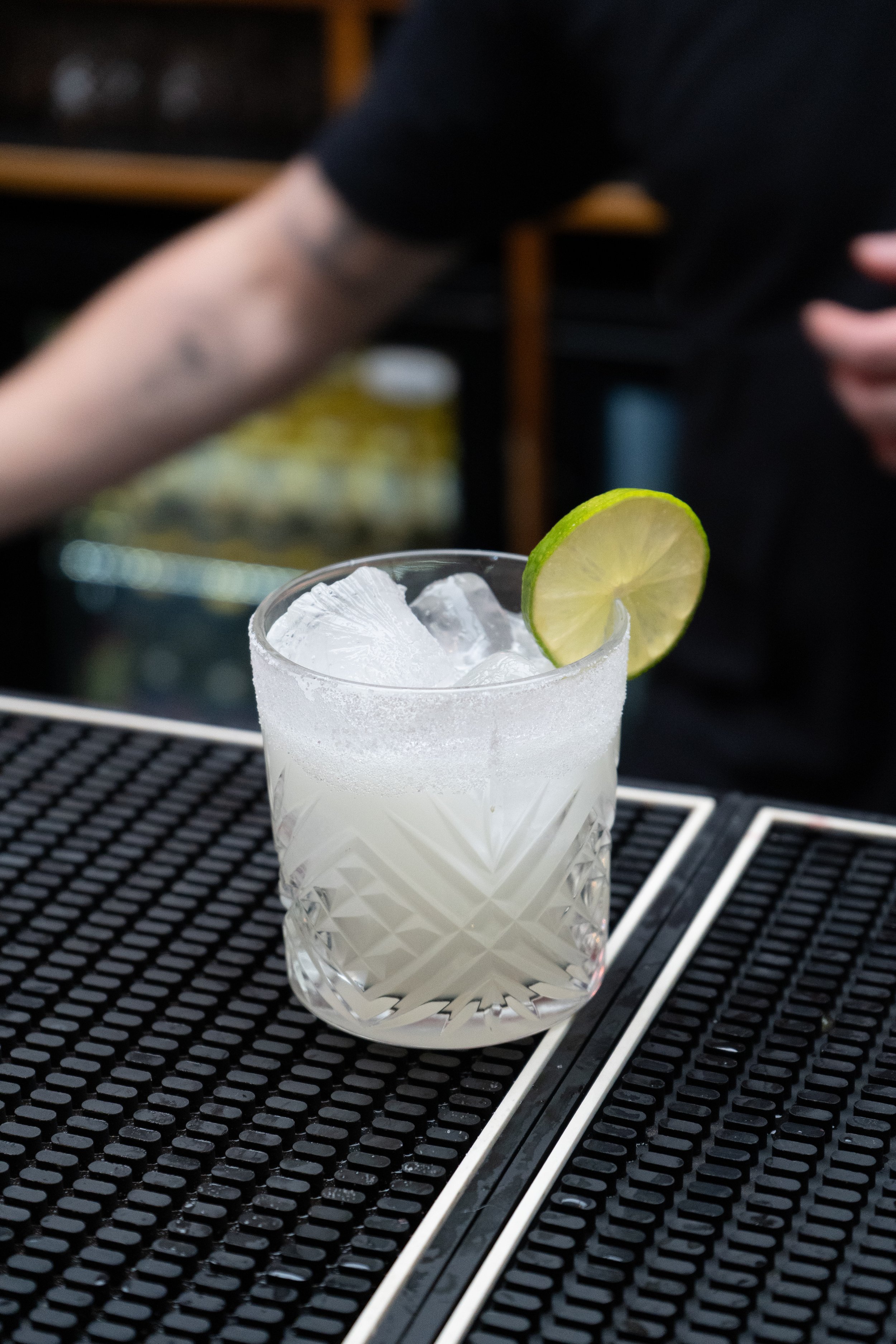 A glass of cocktail with a slice of lime on the rim, filled with ice, sitting on a black rubber bar mat.
